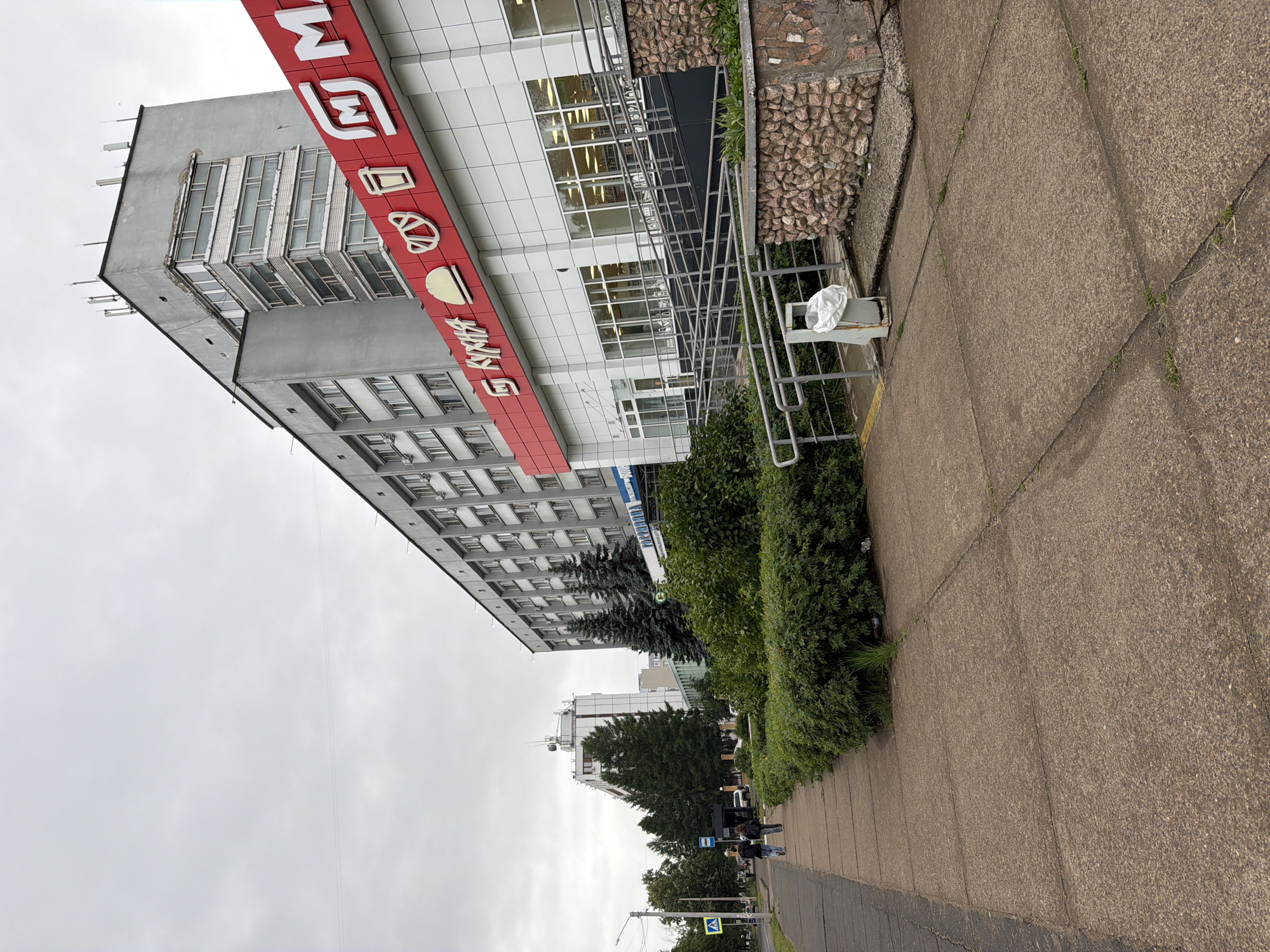 The image shows an outdoor scene in Chaikovsky, Russia. The main subject is a tall, multi-story building with a red and white sign across its facade. The sign has white lettering and graphic symbols, including what appears to be a stylized "M" and images of bread and a coffee cup. Below the sign, the building has rows of windows and a modern white exterior. To the right of the building, there is a walkway or sidewalk made of concrete slabs. Adjacent to the walkway is a dense green hedge. In the background, other buildings and trees are visible under an overcast sky, suggesting cloudy weather.
On the sidewalk, a few people are walking away from the camera. One of them appears to be wearing a dark jacket and carrying a bag. There are also traffic signs visible further down the street, including a blue and white pedestrian crossing sign. The overall mood of the image is neutral, depicting a typical urban environment on a cloudy day. The presence of the red and white sign with food-related imagery on the building suggests it might be a commercial establishment like a cafe or bakery.