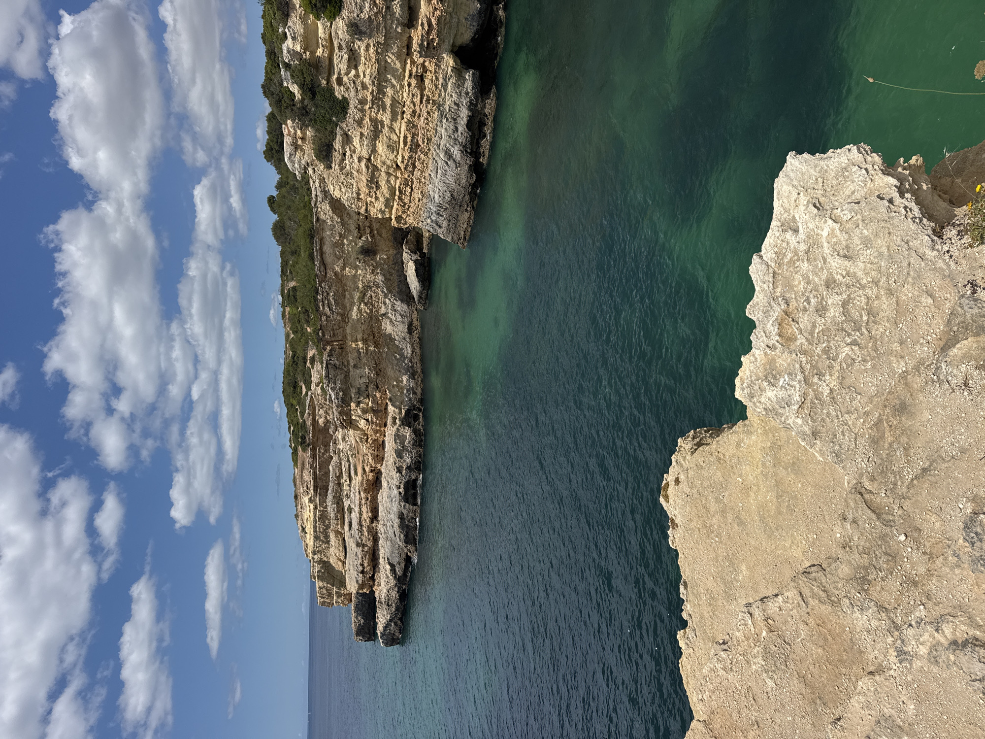 The image depicts a coastal scene with prominent cliffs overlooking a body of water. The cliffs are layered with various shades of brown and tan, displaying geological formations and vegetation. The water appears green and blue. The sky is blue with white clouds. The location is Unknown, Unknown.
