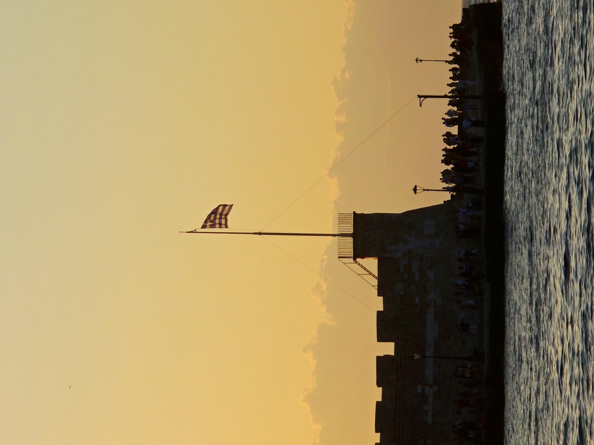 The image captures a silhouette of the Venetian Harbor in Chania, Greece, at sunset. A large stone fortress wall, likely part of the historical sea defenses, forms the dominant structure in the mid-ground. A Greek flag flies prominently from a flagpole atop a circular tower on the fortress. Silhouettes of numerous people are visible walking along the pier and the ramparts of the fortress, suggesting a popular gathering spot. Several lampposts are also visible along the pier. The sky is a warm gradient of orange and yellow, with soft clouds towards the horizon. The sea in the foreground reflects the twilight colors, with gentle ripples on its surface. A single bird is visible in flight in the upper left quadrant of the sky.