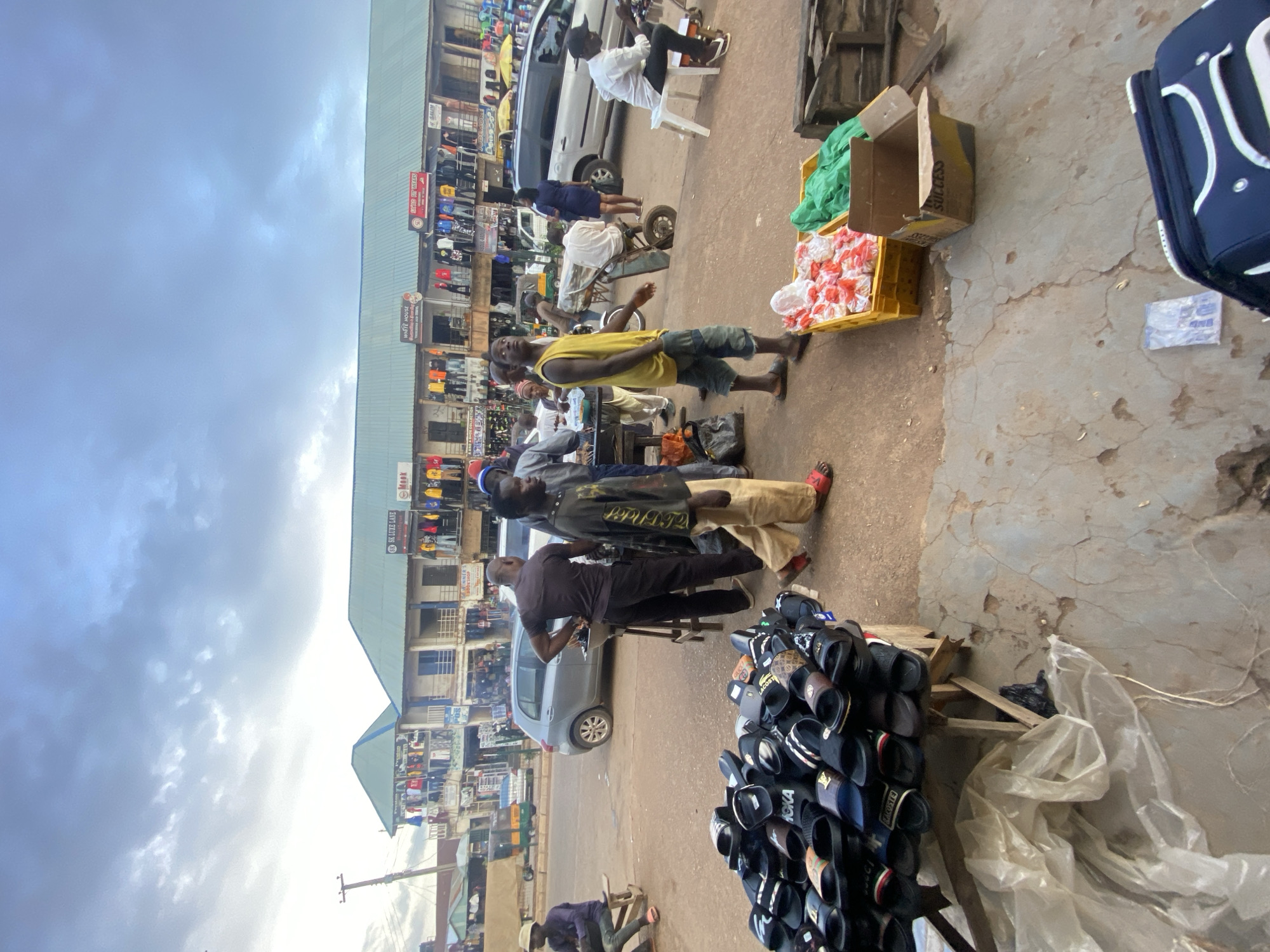 The image depicts a busy street market scene in Bukuru, Nigeria. In the foreground, a stall displays a large collection of black and patterned slide sandals. Adjacent to the sandal stall, two yellow crates are filled with red and white items, possibly packaged goods, and a green material. A brown cardboard box labeled "SUCCESS" sits nearby. A dark blue and white suitcase is partially visible in the lower right corner.
In the mid-ground, several people are engaged in various activities. A man in a yellow tank top and denim shorts stands looking away from the camera. Another man in light-colored pants and a dark shirt is walking away. A man in a dark shirt stands near a cart with a metal frame and wheels, appearing to interact with someone off-camera. A man in a white shirt is seated on a white plastic chair on the right, looking at something in his hands. Further back, a silver car is parked on the street.
The background features a two-story building housing multiple shops. Signs above the shops indicate various businesses, including "SK LUXE LANE", "MOOK", "WHITE HOUSE", and "KING OF WEARS". Clothing, including t-shirts and jackets in various colors, is displayed on racks outside the shops. A three-wheeled auto-rickshaw is visible on the road. The sky is overcast with cloudy conditions. The ground is paved with concrete and dirt.