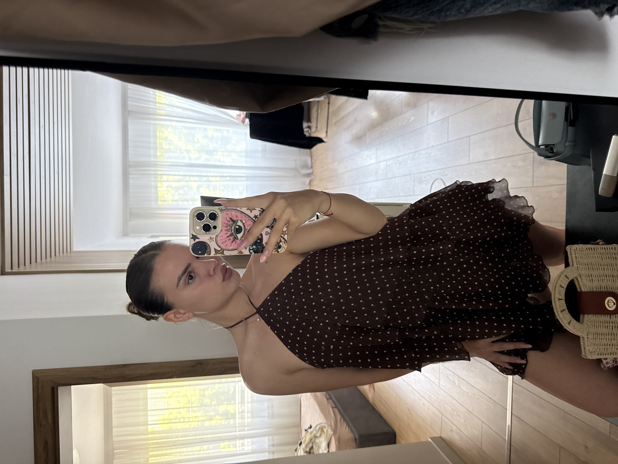 A woman is taking a selfie in front of a mirror. She has her hair pulled back, and is wearing a brown, polka-dot dress. Her phone is in her hand, and it has a decorative case. The mirror reflects a room with wooden floors, blinds on the window, and a bag hanging on the wall. The location is Unknown, Unknown.
