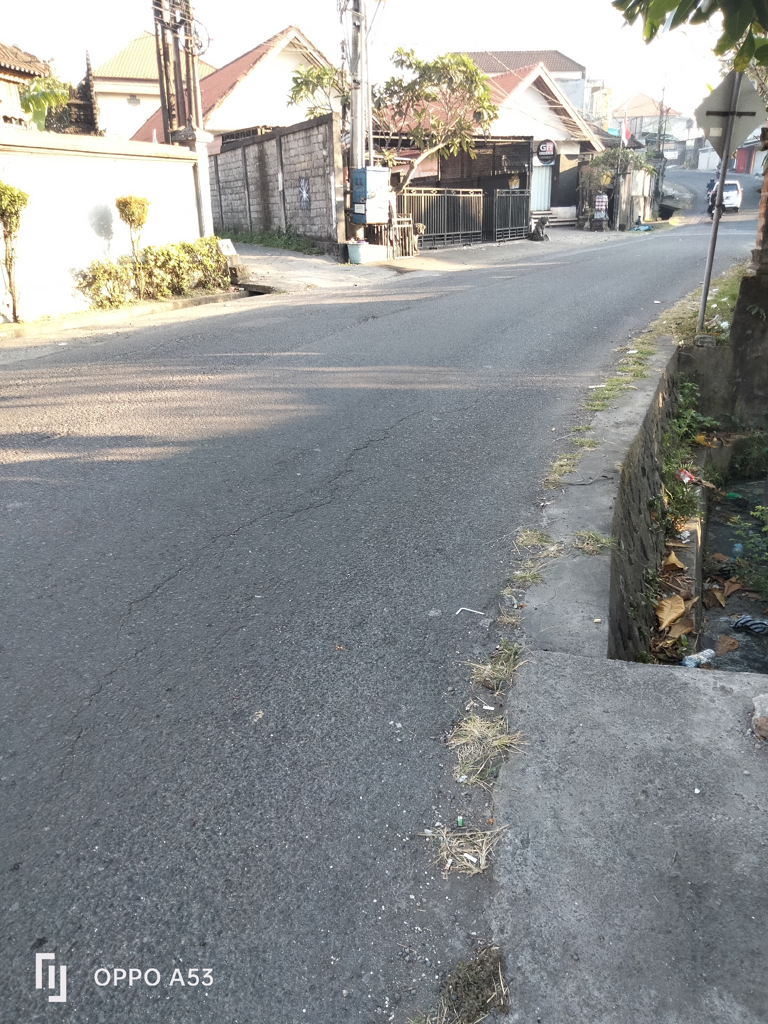 The image depicts a street scene in Denpasar, Indonesia, likely during the morning or late afternoon due to the long shadows cast by the trees. The road is paved and appears to be in moderate condition, with visible cracks. On the left side of the road, there is a white wall, possibly part of a residential property, with a small, well-maintained hedge and two decorative bushes. Further along, there's a brick wall. On the right side of the road, a concrete drain runs alongside the asphalt. This drain contains stagnant water with visible debris, including plastic bottles and dry leaves. The area shows signs of general urban clutter, with scattered trash on the road and in the drain.
In the background, several buildings are visible, including houses with terracotta roofs and some commercial establishments. One building has a sign that appears to read "GR" in black letters on a white background, suggesting a business. A black metal gate is in front of this establishment. A white car is parked or driving on the road in the distance, and there are utility poles with electrical wires overhead. The overall atmosphere is that of a typical urban street in a tropical climate, with vegetation and sunlight playing a significant role in the visual composition. The watermark "OPPO A53" is present in the bottom left corner, indicating the device used to capture the image.
