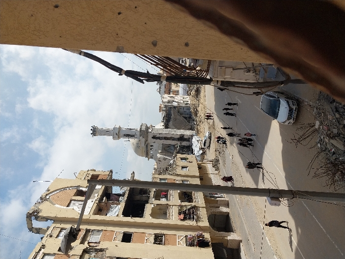 This image depicts a damaged urban environment in Khan Yunis, Palestinian Territory, under a partly cloudy sky.
In the foreground on the right, the light-colored, pockmarked wall of a building is visible, with multiple dark utility cables running vertically down its side. Below this wall, a pile of concrete rubble and exposed rebar lies at the edge of a paved street.
The street, a two-lane asphalt road, shows areas of light-colored dust or debris. A white Hyundai Tucson SUV is parked facing away from the viewer on the right side of the street. Approximately 15 individuals, including adults and children, are walking on the street. A lone person in dark clothing walks on the left side of the street in the foreground. Several small groups of people are scattered along the road, generally moving away from the viewer. Further down the street, a light-colored sedan is visible near a large debris pile.
To the left, a multi-story building (at least five floors visible) exhibits extensive damage, with large sections of its facade missing, exposed interiors, and numerous holes consistent with projectile impacts. Clothes are hanging from multiple balconies or window openings. A concrete utility pole with a street lamp is positioned in front of this building.
In the mid-ground, a prominent mosque features a light-colored dome and a tall, slender minaret, which appears damaged near its top section. The structures immediately surrounding the mosque are heavily damaged or reduced to rubble. Beyond the mosque, further into the background, additional damaged buildings and debris piles are visible.