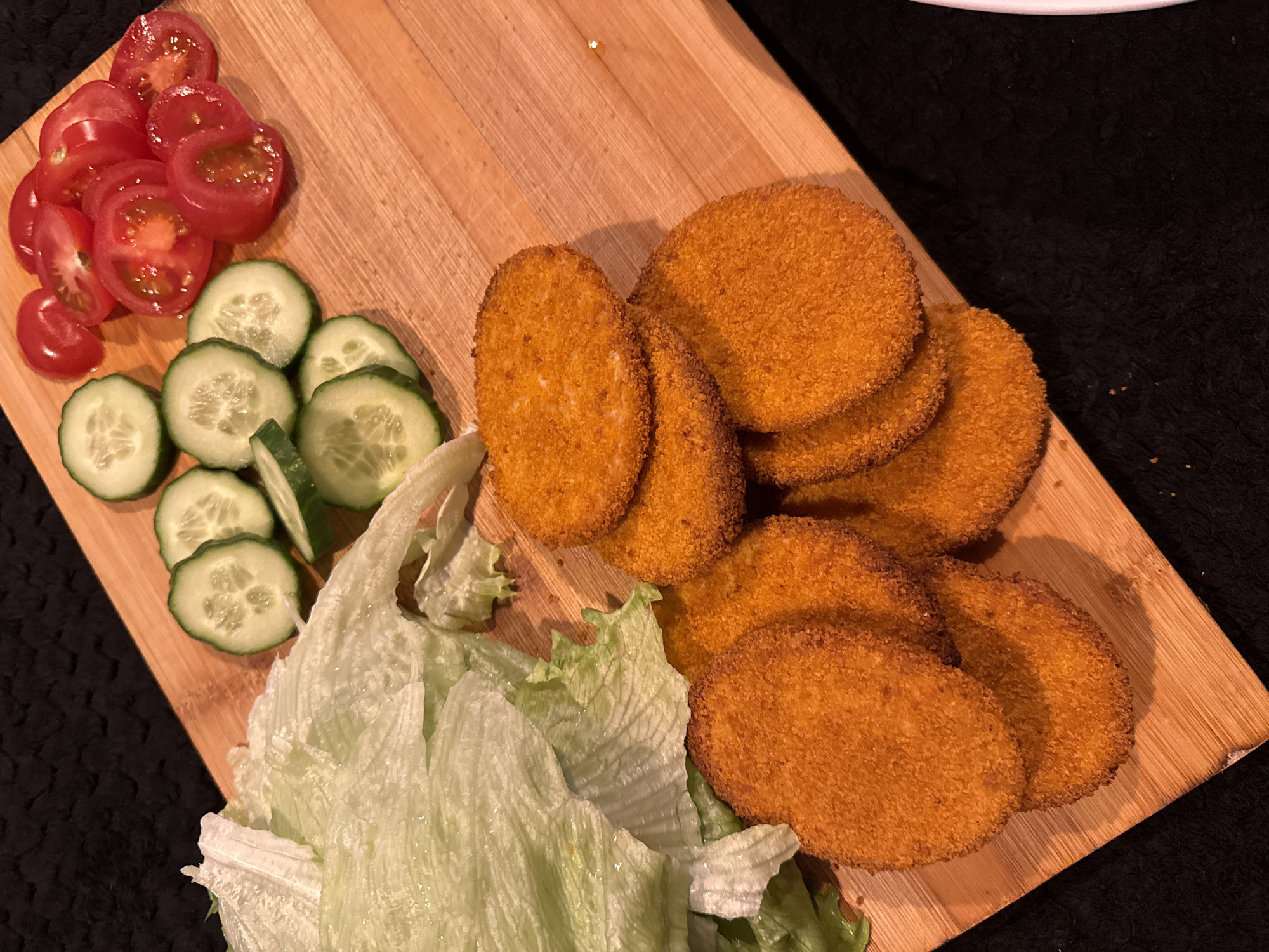 The image shows a wooden cutting board on a black surface. The cutting board holds sliced cherry tomatoes, sliced cucumbers, lettuce, and a stack of fried, round food items. The image does not contain information about Great Yarmouth or any other city. There are no people present in the image.