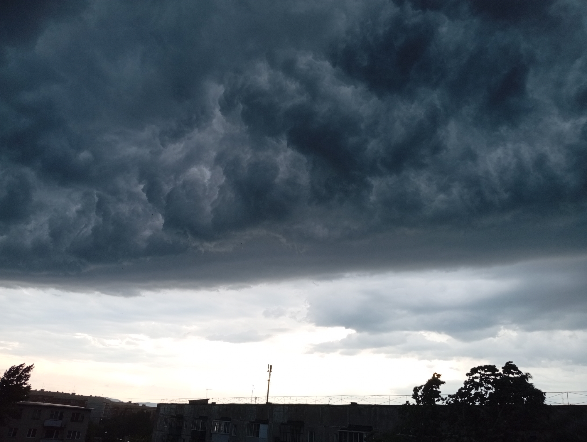 The image depicts a dramatic sky filled with dark, heavy storm clouds looming over the city of Kyshtym, Russia. The clouds occupy the upper two-thirds of the frame, their textured surfaces suggesting an impending downpour. Below the storm clouds, a lighter band of sky breaks through, revealing the softer hues of twilight. In the foreground, the silhouette of buildings and trees is visible against this lighter sky. The architectural style of the buildings suggests Soviet-era construction, a common feature in many Russian cities. A cell tower stands prominently on the rooftop of one building, its antenna reaching towards the turbulent sky. The overall impression is one of atmospheric intensity and the quiet presence of urban life beneath a powerful natural display.
