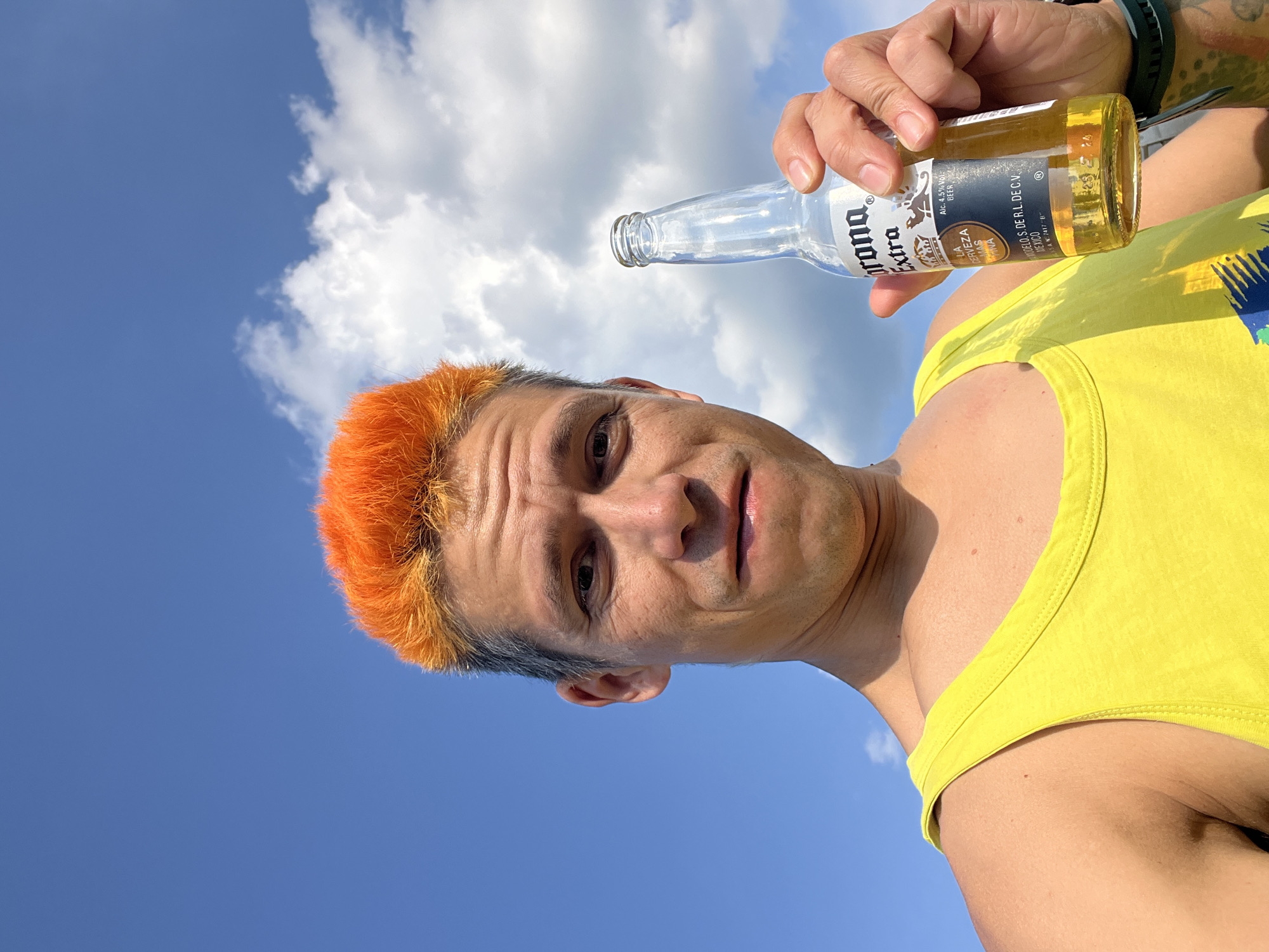 A man with bright orange, spiked hair is holding a bottle of Corona Extra beer and looking at the camera. He is wearing a yellow tank top and is outdoors with a bright blue sky and white clouds in the background. The image is a close-up, focusing on the man's face and upper body. The location is not specified in the image or its metadata.