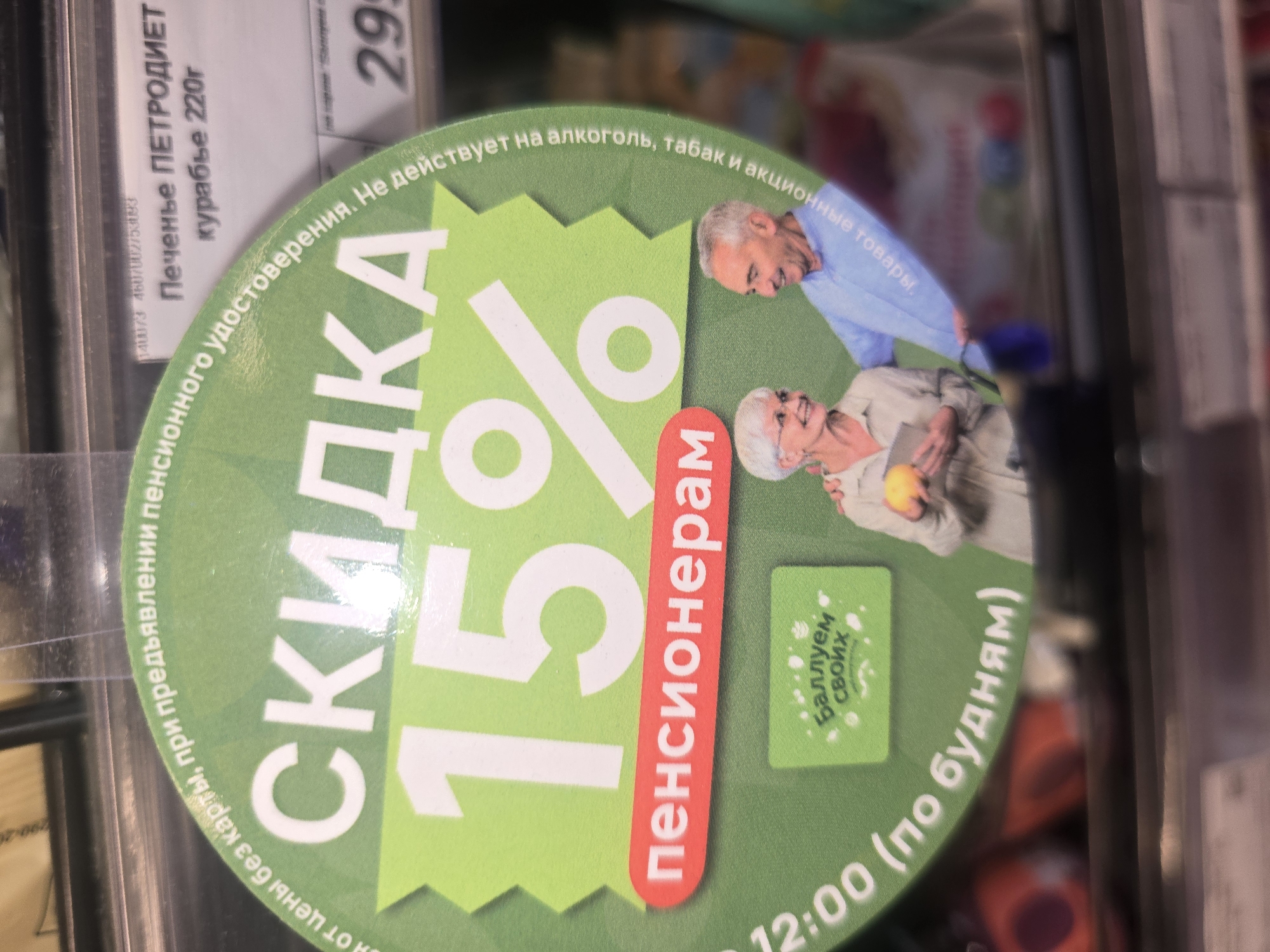 The image shows a close-up of a circular green sticker with text and graphics, likely displayed in a retail store in Ulan-Ude, Russia. The sticker advertises a discount for pensioners.
The main text on the sticker reads "СКИДКА 15%" which translates to "DISCOUNT 15%". Below this, in a red oval, is the word "пенсионерам", meaning "for pensioners". The sticker also states that the discount is "без карты, при предъявлении пенсионного удостоверения" which means "without a card, upon presentation of a pension certificate".
In the background of the sticker, two elderly individuals, a woman and a man, are depicted smiling. The woman is wearing glasses and a light-colored shirt, holding a book. The man is wearing a blue shirt and has his arm around the woman.
Around the edge of the green sticker, more text is visible, including "действует на алкоголь, табак и акционные товары" which translates to "valid on alcohol, tobacco, and promotional items". There's also text that appears to be "12:00 (по будням)" meaning "12:00 (on weekdays)".
To the right of the sticker, a price tag is partially visible with the number "299" and a product name that appears to be "Печенье ПЕТРОДИЕТ" (Petrodiet Cookies) and "куратье 220г" (220g kurabye).
The overall environment suggests a supermarket or a similar retail setting in Ulan-Ude, Russia. The lighting and background indicate the sticker is attached to a shelf or display.