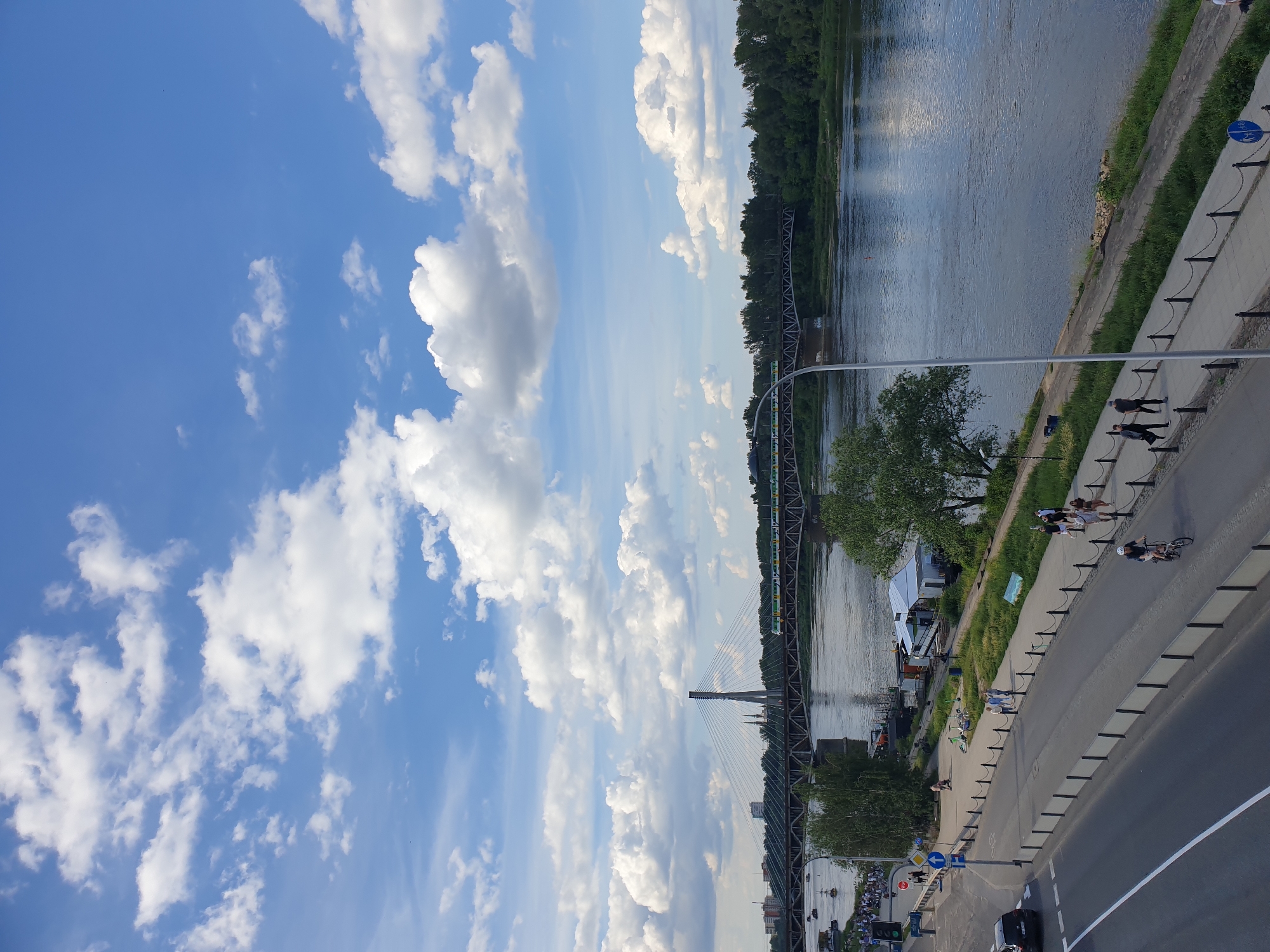 The image presents an elevated view overlooking the Vistula River in Warsaw, Poland, under a bright blue sky scattered with white cumulus clouds.
In the mid-ground, the cable-stayed Świętokrzyski Bridge spans the river, characterized by its distinctive single pylon on the left bank. Behind it, the steel truss structure of the Most Średnicowy (Średnicowy Bridge), a railway bridge, is visible, with a green and yellow train actively crossing it from right to left. The city skyline is faintly discernible in the distance behind the bridges. The far bank of the river is densely covered in trees and vegetation. Several small boats or barges are present on the river, predominantly on the left side near the bank.
In the foreground and lower mid-ground, the left bank of the river features a multi-lane road and a parallel pedestrian promenade. A black car is visible on the road in the lower left. Numerous individuals are engaged in various activities on the promenade and adjacent sidewalks: several people are walking in groups or individually, and a person wearing a helmet is cycling on a path near the riverbank on the lower right. The promenade is bordered by a chain-link fence system and intermittent trees. Small structures, possibly boat sheds or kiosks, are situated close to the water's edge on the left bank. A tall lamppost extends from the bottom center, partially obscuring the view of the river and far bank.