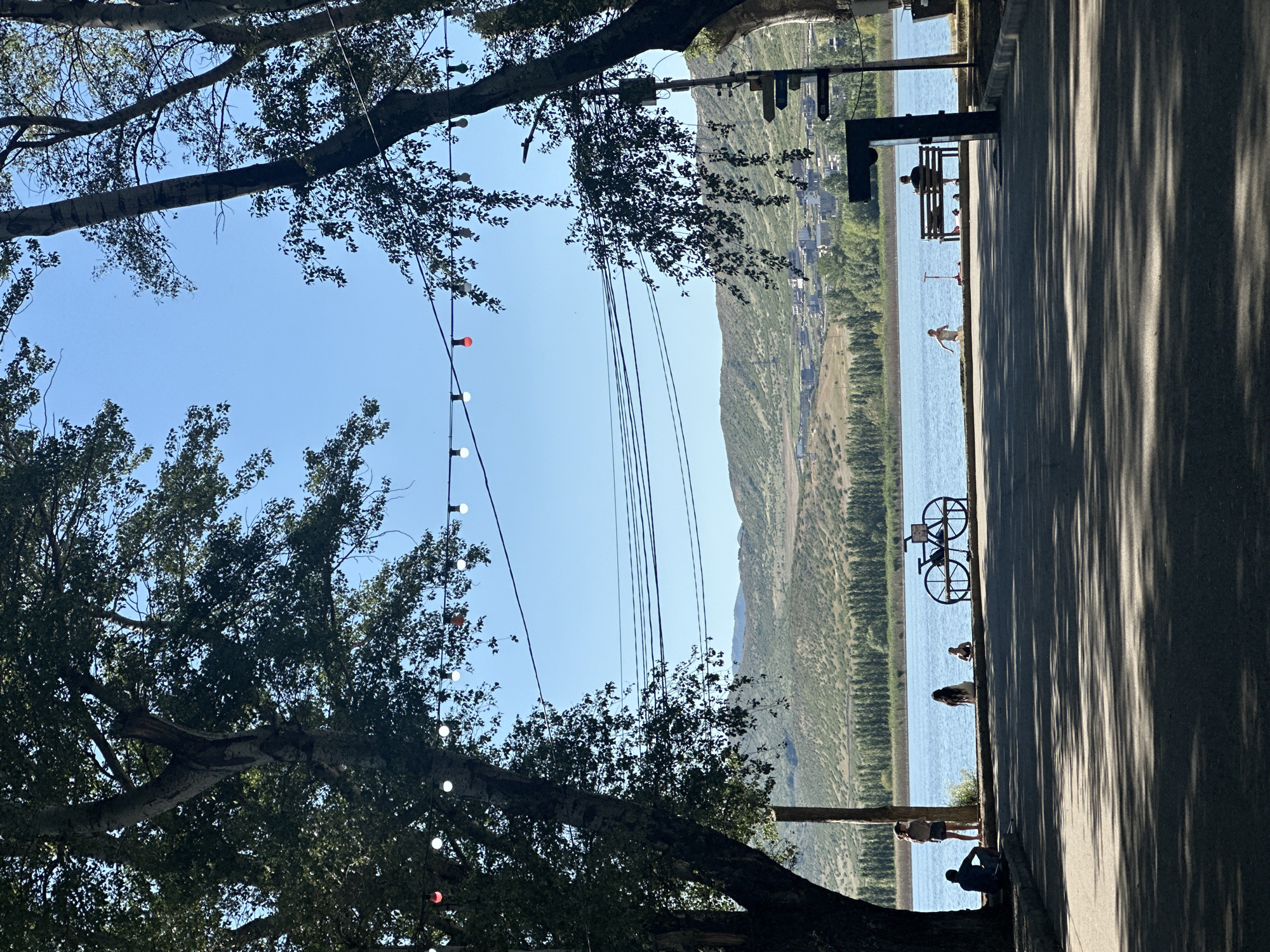 This image captures a serene waterside scene, likely near Tbilisi, Georgia, on a clear, sunny day. In the foreground, a paved pathway is partially shaded by the dappled sunlight filtering through the leaves of mature trees. Beyond the pathway, a body of water, possibly a lake or a wide river, stretches across the mid-ground, its surface reflecting the bright blue sky.
Along the edge of the water, a few individuals are visible. On the left side, two people are seated on the ground under the shade of a large tree. Further along the waterfront, three people are standing near the water's edge. To their right, a decorative black metal bicycle structure is placed, serving as a potential bike rack or ornament.
In the distance, across the water, a landscape of rolling hills covered in scrub and trees rises, dotted with scattered buildings and a dense line of trees along the lower slopes. The sky is a vibrant, cloudless blue, and strung across the upper portion of the image is a string of decorative lights, some illuminated with red and white bulbs. On the right side of the frame, a modern-looking dark structure with directional signs is positioned near the waterfront. A person is seated on a park bench facing the water, while further down the path, a child is running and a red scooter lies on the ground. The overall atmosphere is one of peaceful recreation and natural beauty.