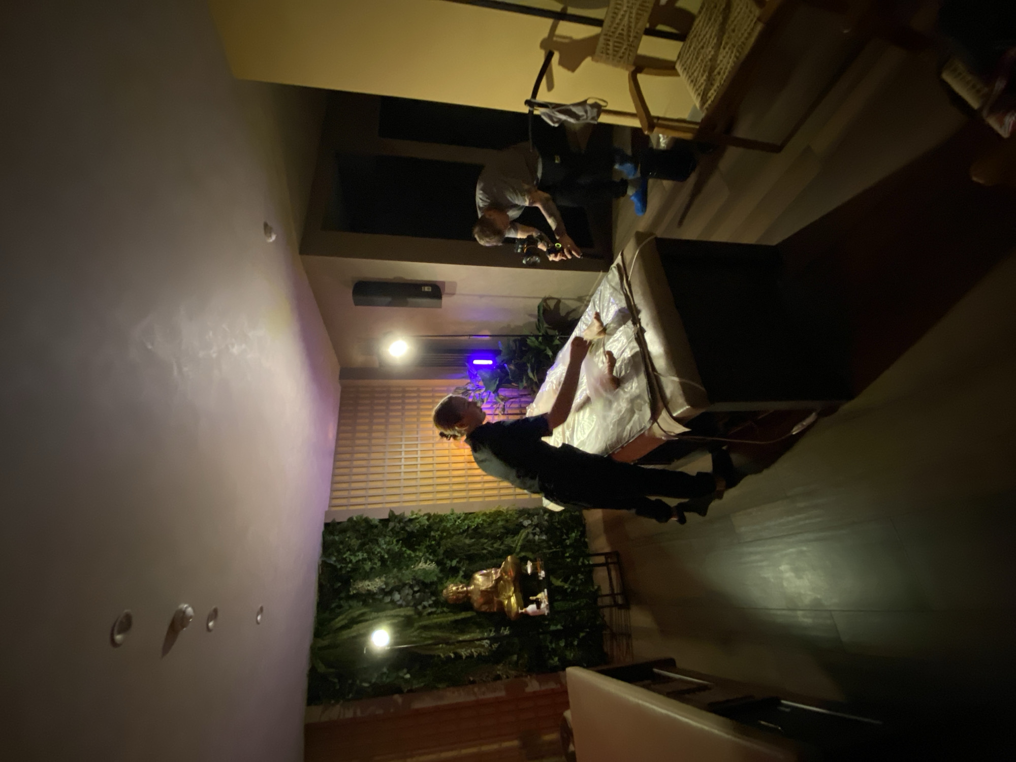 The image shows an indoor scene with two people, likely in a commercial establishment. One person is wearing a dark shirt and pants and appears to be standing near a table covered with a light-colored cloth. The second person is wearing a light-colored shirt and dark pants and is crouching, possibly operating a camera or some other device. In the background is a green wall with what appears to be a golden statue. Near the top of the image are chairs and some equipment. The lighting is somewhat dim, with a mix of artificial and natural sources. While it's not possible to determine the location in Izhevsk, Russia, based on the image alone, the details provided are comprehensive for an objective assessment of the scene.