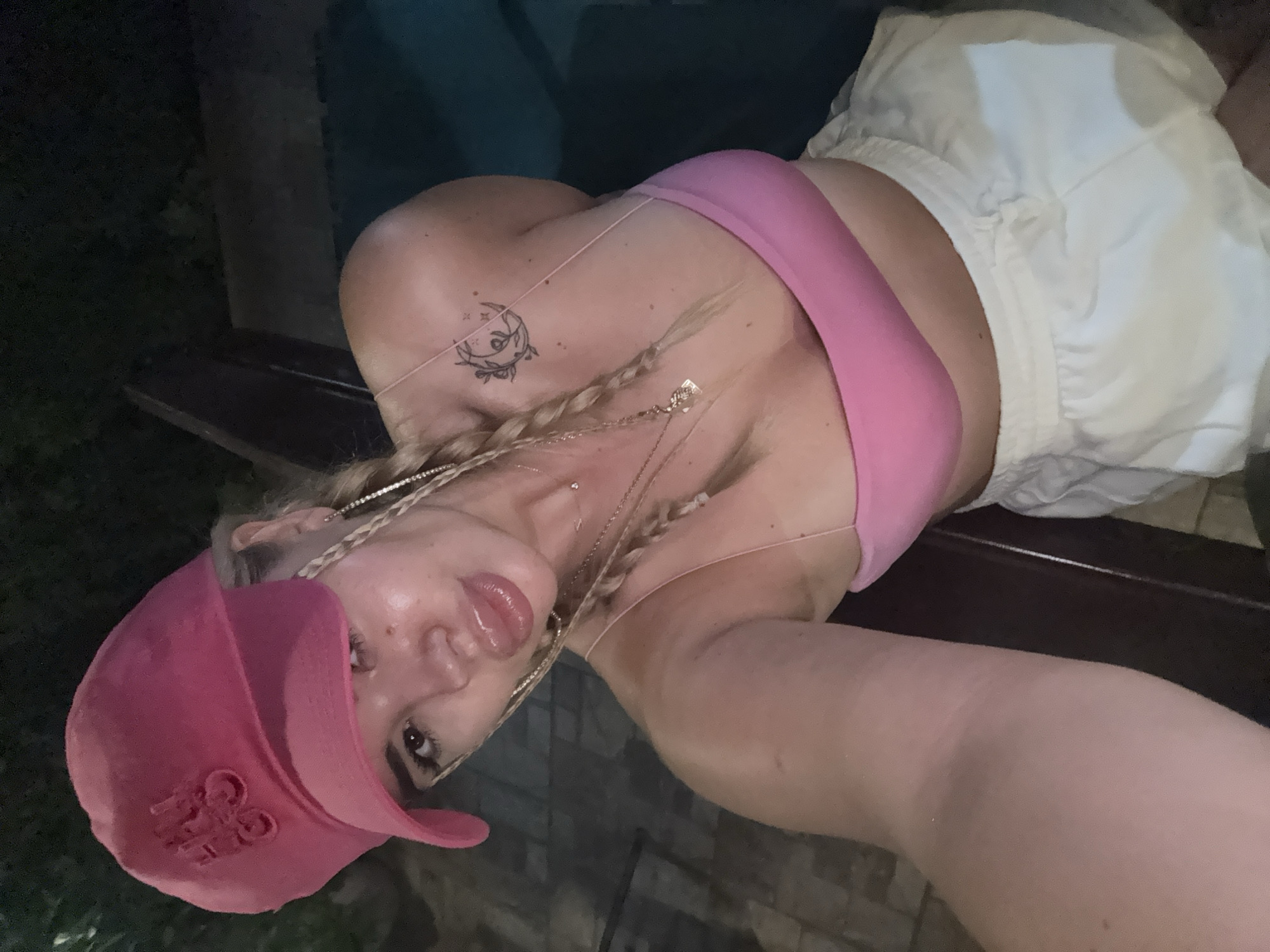 A young woman with blonde hair in braids, wearing a pink baseball cap and a pink bikini top with white shorts, is taking a selfie outdoors at night. She has a crescent moon tattoo on her left shoulder. The background suggests an urban setting with some greenery and possibly a pool. The image does not provide any specific details about the city of Rakovski, Bulgaria.