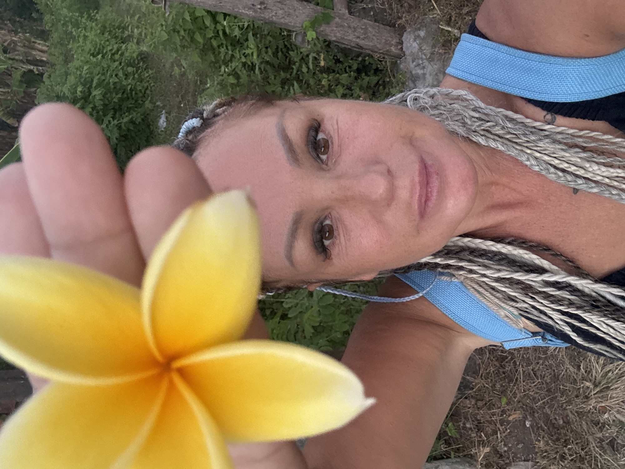 A woman with blonde braids stands outdoors in Kuta, Indonesia. She is holding a yellow flower in front of her face. The woman is wearing a blue tank top. The background consists of green foliage and a tree.