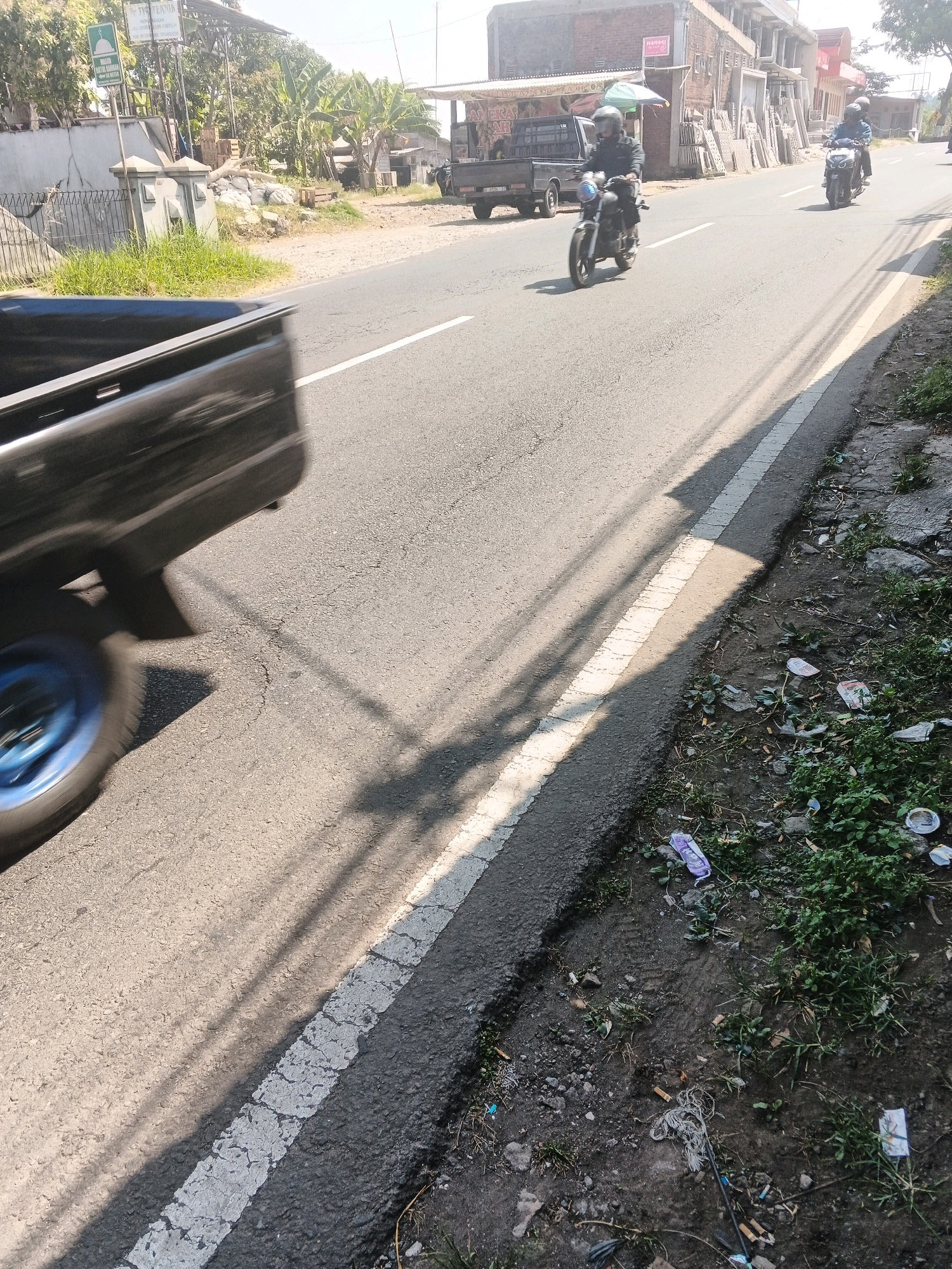 The image depicts a street scene in Bandungan, Indonesia. In the foreground, the blurry blue wheel and rear section of a black pickup truck are visible, indicating motion. The truck is on the right side of the road, moving away from the viewer. The road is asphalt, with a white line marking the edge of the lane.
In the middle ground, two motorcycles are traveling in the same direction as the pickup truck. The first motorcycle, closer to the viewer and slightly to the left, is ridden by a person wearing a helmet and dark clothing. The second motorcycle, further down the road and to the right, is also ridden by a person wearing a helmet.
The background shows buildings and trees. The buildings appear to be commercial establishments, with some materials stacked outside. There are also banana trees and other vegetation, typical of a tropical environment. The sky is clear and bright, suggesting a sunny day. The roadside on the right is unpaved and shows signs of litter, including plastic wrappers and cigarette butts. Shadows of trees and possibly power lines are cast across the road, indicating the direction of sunlight.