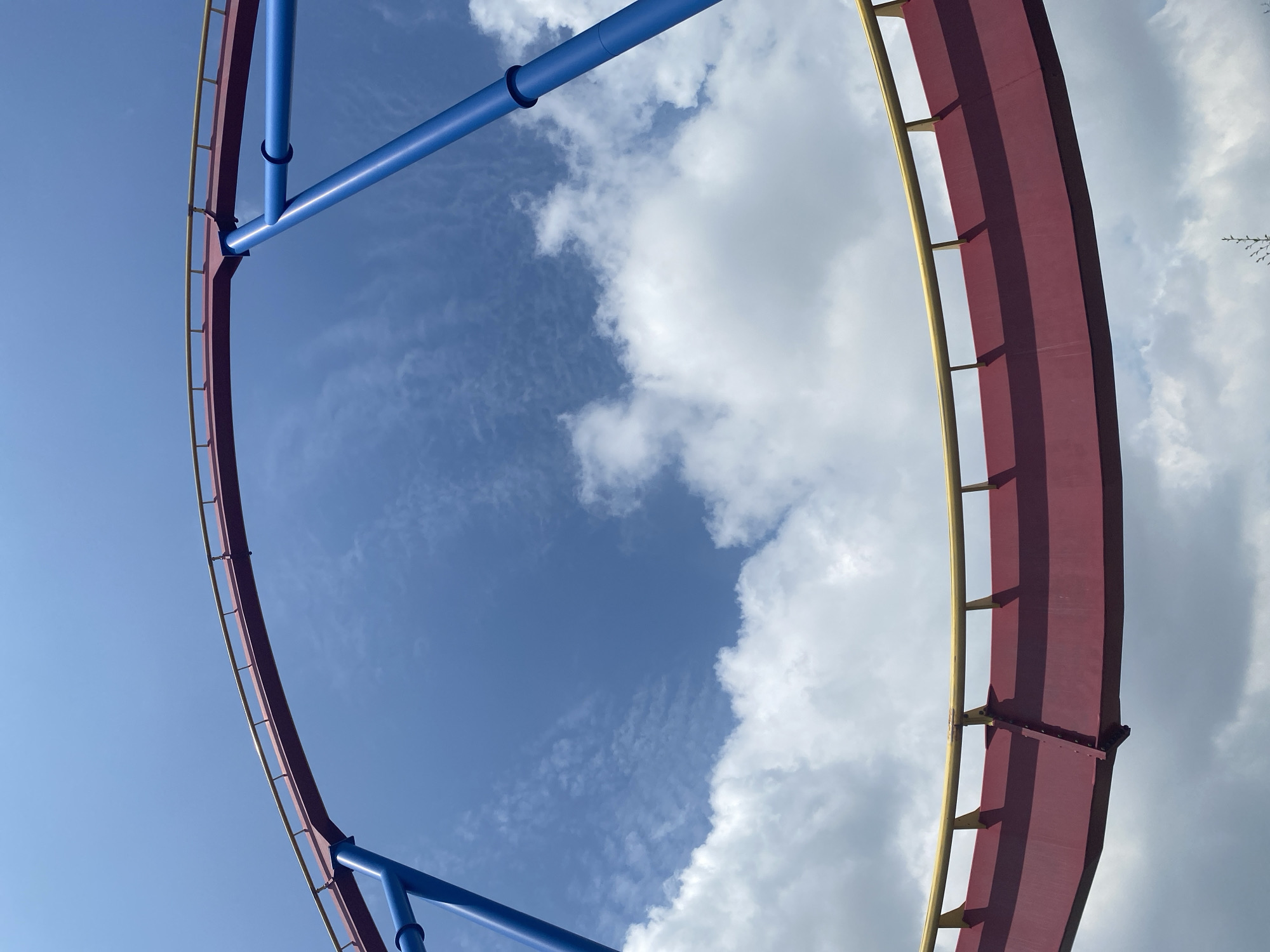 The image shows a section of a roller coaster track against a blue sky with scattered white clouds. The track is constructed with red supports and a yellow railing. Two bright blue poles extend upwards, supporting a curved section of the track. The perspective is from below, looking up at the structure. The scene is outdoors, suggesting an amusement park or similar recreational facility. The provided crops focus on different parts of the roller coaster and the sky. No specific location in Montreal, Canada can be identified from the image.