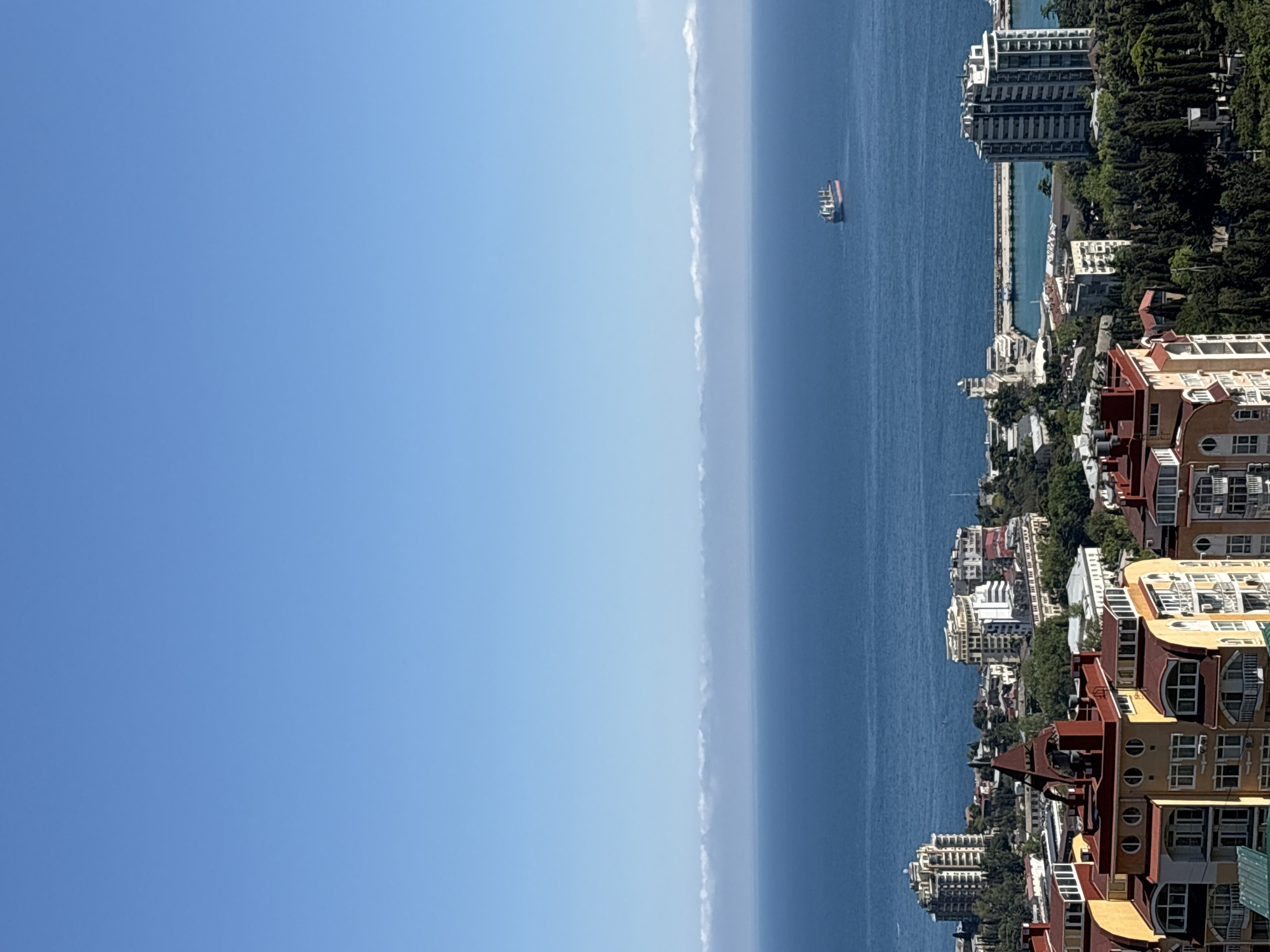 The image depicts a vertical, panoramic view of a coastal city, identified as город Ялта (Yalta), Ukraine. A clear, blue sky dominates the left half of the frame, transitioning into a lighter haze near the horizon. A sliver of the sea lies in the middle, with a large cargo ship visible on the water. Along the right side of the image, there is a densely populated coastline featuring a variety of buildings, ranging from high-rise apartments to smaller residential structures, interspersed with patches of greenery and trees. A pier or harbor structure can also be seen. The buildings are diverse in architectural style and color.