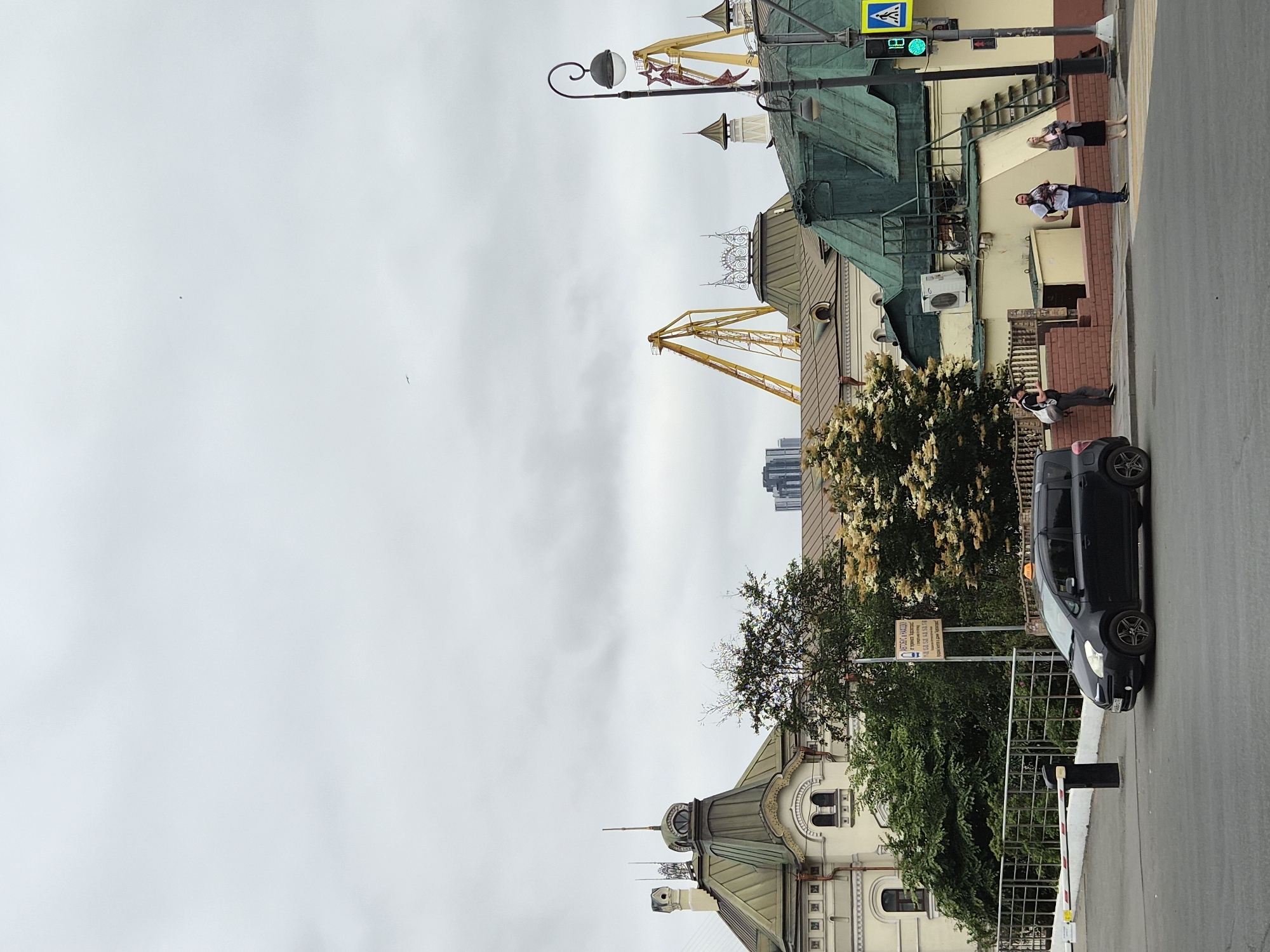 The image captures an overcast day in a city, likely Vladivostok, Russia, given the presence of port cranes in the background. In the foreground, a black Toyota Ractis taxi is stopped at an intersection. To the right, a pedestrian crossing signal shows a green walking figure, indicating it is safe to cross. Three individuals are visible on the sidewalk: one man in a white t-shirt and shorts carrying a backpack, another man in a grey jacket and black skirt, and a third man in a black t-shirt and shorts with a backpack. Behind them, a building with a green roof and a yellow facade stands. Further in the background, several industrial cranes are prominent, suggesting a port or industrial area. Architectural elements of older buildings with distinctive green roofs and turrets are also visible. The sky is heavily clouded.