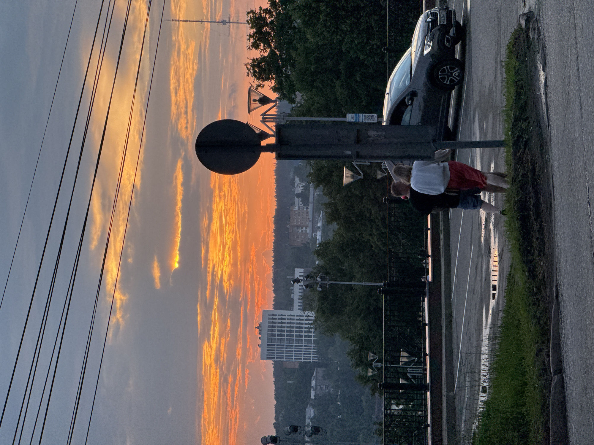 This is a medium-distance, eye-level, outdoor shot taken at dusk in Izhevsk, Russia. The sky is filled with dramatic, orange and gray clouds, suggesting a sunset. In the background, a modern, white, multi-story building with many windows stands out against a backdrop of dark green trees. A tall, lattice radio tower is visible on the right side of the horizon. In the foreground, a gray car is parked in a lot with white markings. Two individuals, a man wearing a dark t-shirt and blue shorts, and a woman in a white top and red shorts, are standing near the car. A dark, circular traffic sign is positioned prominently on a post in the center-right of the frame, obscuring part of the scene. The ground appears wet, with puddles reflecting the sky, indicating recent rain. Power lines are visible crisscrossing the top of the image.