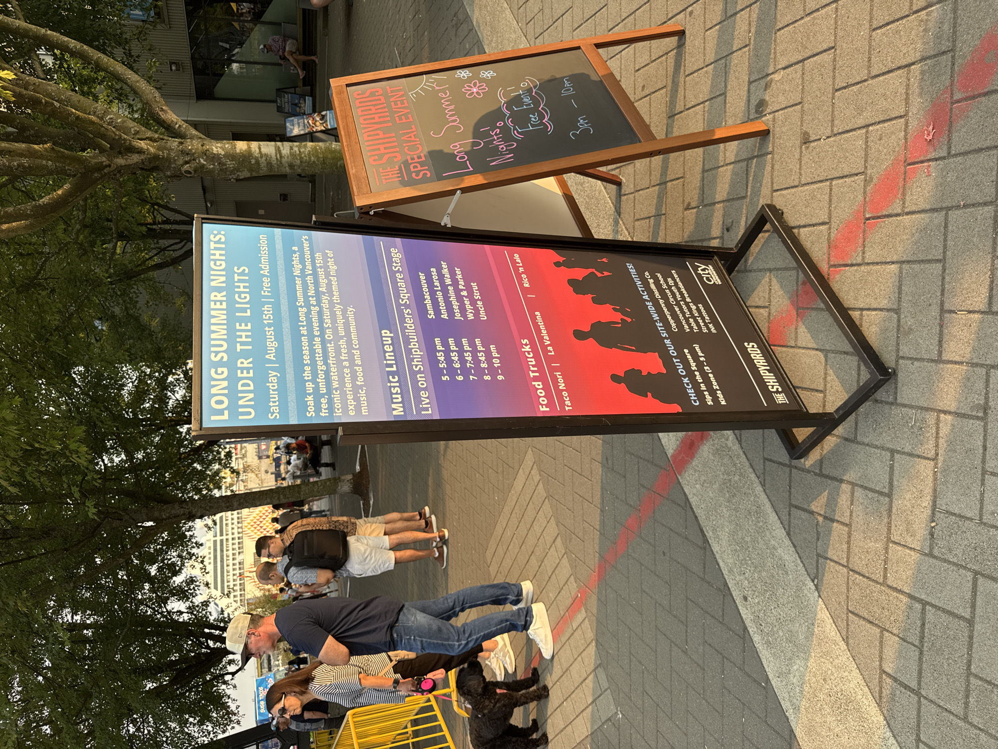 The image shows an outdoor scene at The Shipyards in North Vancouver, Canada, on a sunny day. A black A-frame sign in the foreground displays "THE SHIPYARDS SPECIAL EVENT" and "Long Summer Nights! Free Event!" with "3pm - 10pm" written below. To the left of this sign, a taller, light blue sign lists "LONG SUMMER NIGHTS: UNDER THE LIGHTS" and details about the event, including the date, "Saturday | August 15th | Free Admission," and information about music lineups and food trucks. The background shows trees, buildings, and people mingling. A group of people, including a man in a blue t-shirt and jeans, a woman in a striped shirt, and a man with a backpack, are walking, with a small black dog on a leash in the foreground. Several yellow barriers are visible on the left side of the image. The ground is paved with light-colored bricks.
