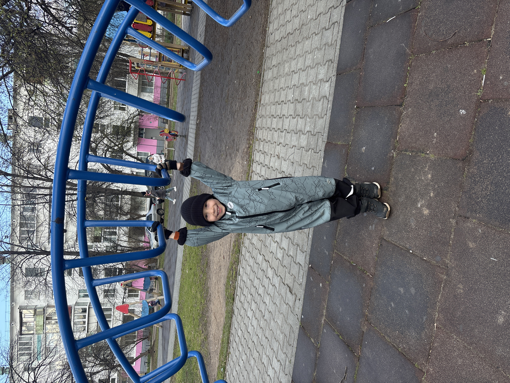 The image depicts a child hanging from a blue metal playground structure in Chaikovsky, Russia. The child, wearing a black beanie and a gray and black patterned jumpsuit, is smiling and holding onto the structure with both hands. The playground is located in an urban environment, with apartment buildings and bare trees visible in the background. The ground is covered in paving stones and patches of grass. Additional playground equipment can be seen in the background.