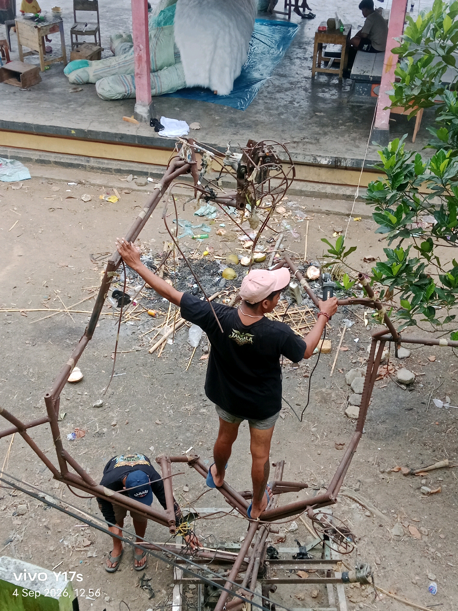 An overhead shot captures two men working on a complex metal structure in an outdoor, cluttered environment. The scene appears to be in Rama Dewa, Indonesia, as indicated by the timestamp "4 Sep 2026, 12.56" and the text "vivo Y17s" which is a phone model.
One man, wearing a pink cap and a black t-shirt with "JANGALA" printed on it, is standing on the metal structure, holding an angle grinder and actively cutting a metal pipe. He is barefoot.
Another man, wearing a blue baseball cap and a black t-shirt, is positioned lower on the structure, bent over and attending to some wires. He is wearing flip-flops.
The metal structure itself is intricate and appears to be in various stages of construction or repair, with exposed pipes, joints, and some attached wires. It is supported by scaffolding-like elements and some wooden planks.
The surrounding ground is unpaved and dusty, with scattered debris including dry leaves, broken coconut shells, plastic bags, and various pieces of metal and wood. In the background, there are some large, white, fluffy objects, possibly part of a decoration or art installation, and some bundled items covered in plastic sheeting. There are also wooden chairs and tables in the background, suggesting a workshop or storage area. The overall impression is one of an informal, working outdoor space.