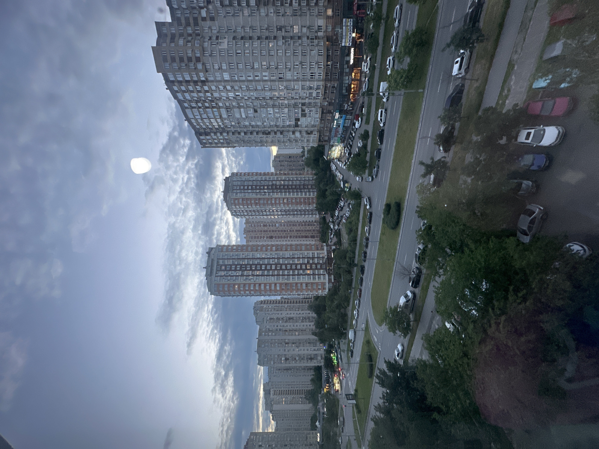 The image depicts an urban landscape in Kyiv, Ukraine, during twilight. Dominating the scene are several tall residential buildings, characteristic of Soviet-era architecture, with numerous windows and balconies. The sky is overcast with diffused clouds, and a bright, circular object, possibly the moon or a light artifact, is visible in the upper portion of the sky. A wide, multi-lane road with a divided median runs through the foreground, with cars parked along its sides and in designated lots. Trees with green foliage line the road and are interspersed between the buildings. The streetlights are beginning to illuminate, casting a soft glow on the asphalt. The overall impression is one of a dense urban environment at the end of the day.