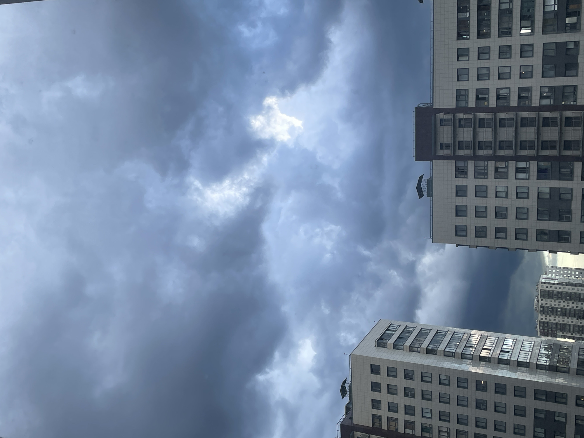The image shows a view of several modern apartment buildings against a dramatic, cloudy sky. The clouds are dark and ominous, with patches of lighter sky peeking through in the upper right quadrant. The buildings are tall and rectangular, with many windows. The building on the left is partially obscured by the dark clouds. The building on the right is more fully visible and features balconies. In the background, further apartment buildings are visible. The scene appears to be urban.