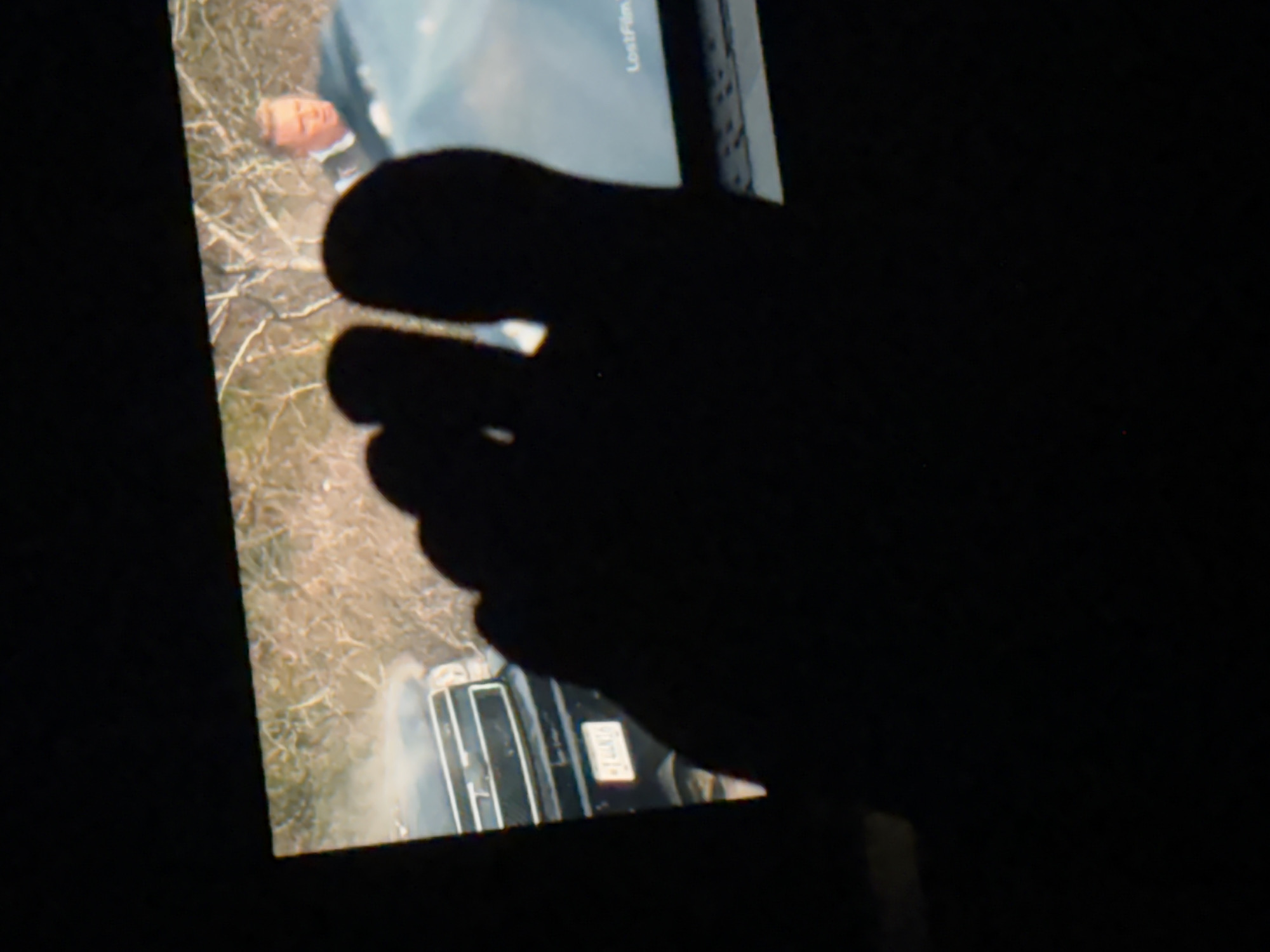 A dimly lit, partially obscured image shows a screen displaying a scene involving a man lying on the ground near a vehicle. A dark shadow, likely a person's foot, covers a significant portion of the screen, obscuring details. The location and specific details of Budva, Montenegro, are not discernible from the image.
