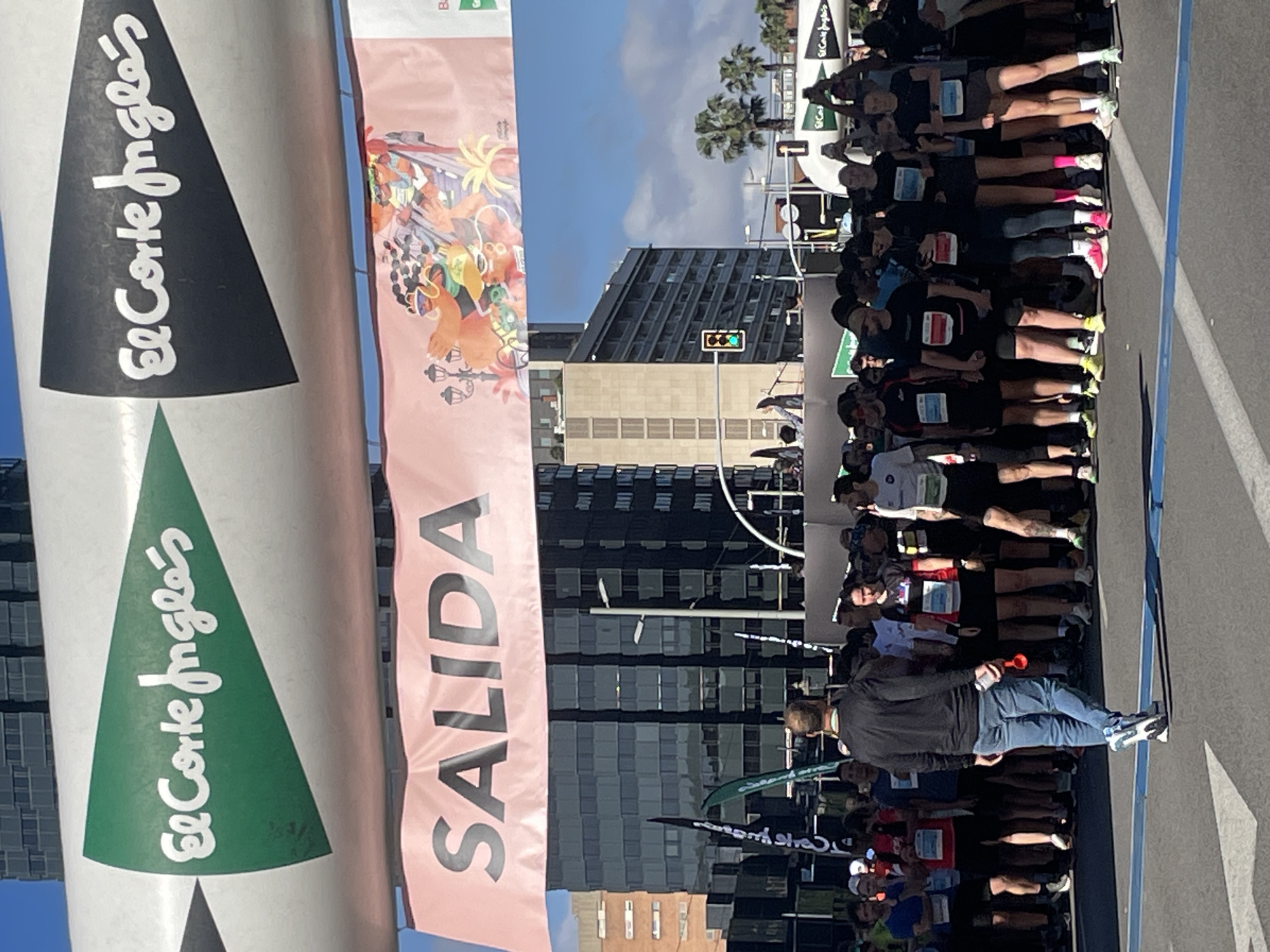 The image depicts the start of a race in Barcelona, Spain, under a clear sky with a modern building and palm trees in the background. Runners are lined up at the starting line, identifiable by the blue marking on the pavement. The "SALIDA" banner, signaling the start, is visible alongside "El Corte Inglés" branding on pillars, identifying a sponsorship. A man in a black jacket and jeans stands out within the crowd of runners, suggesting he could be an organizer or spectator. The scene implies a public event, likely a marathon or similar race, in an urban environment.