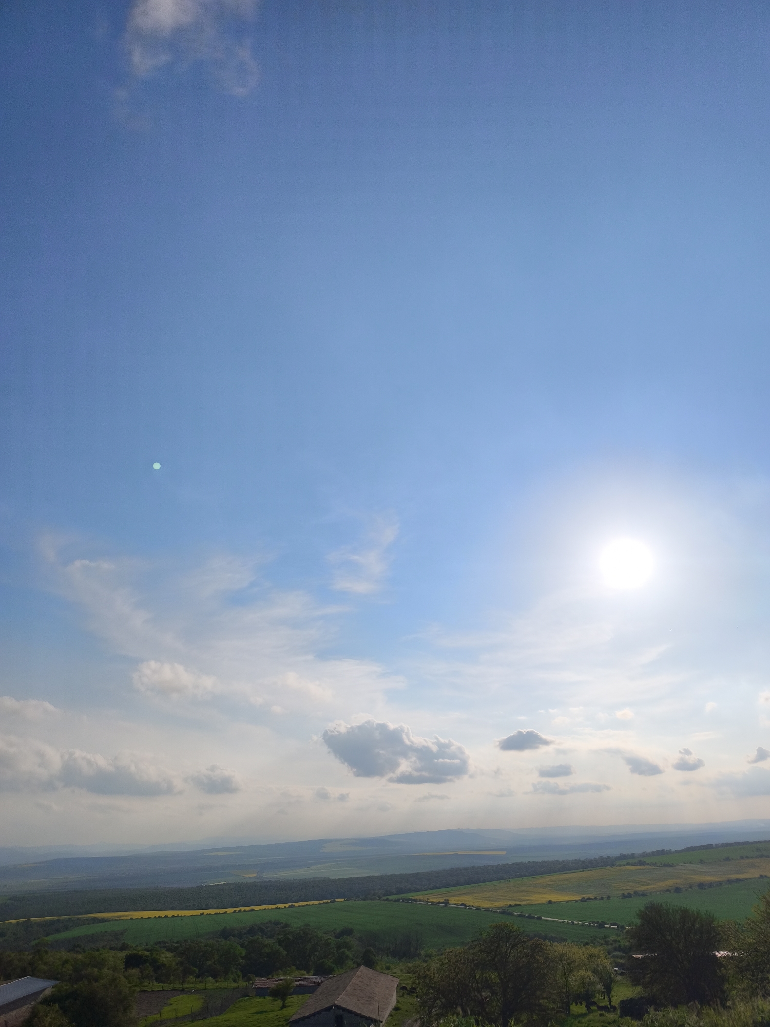 The image depicts a landscape in Avren, Bulgaria, under a bright, sunny sky with scattered clouds. The foreground features a rural scene with greenery and a few buildings with dark roofs. Beyond that, a patchwork of green and yellow fields stretches into the distance, suggesting agricultural land. In the background, rolling hills and a hazy horizon fade into the blue sky. The sun is positioned high in the sky, creating a bright glare. There are no people visible in the image.
