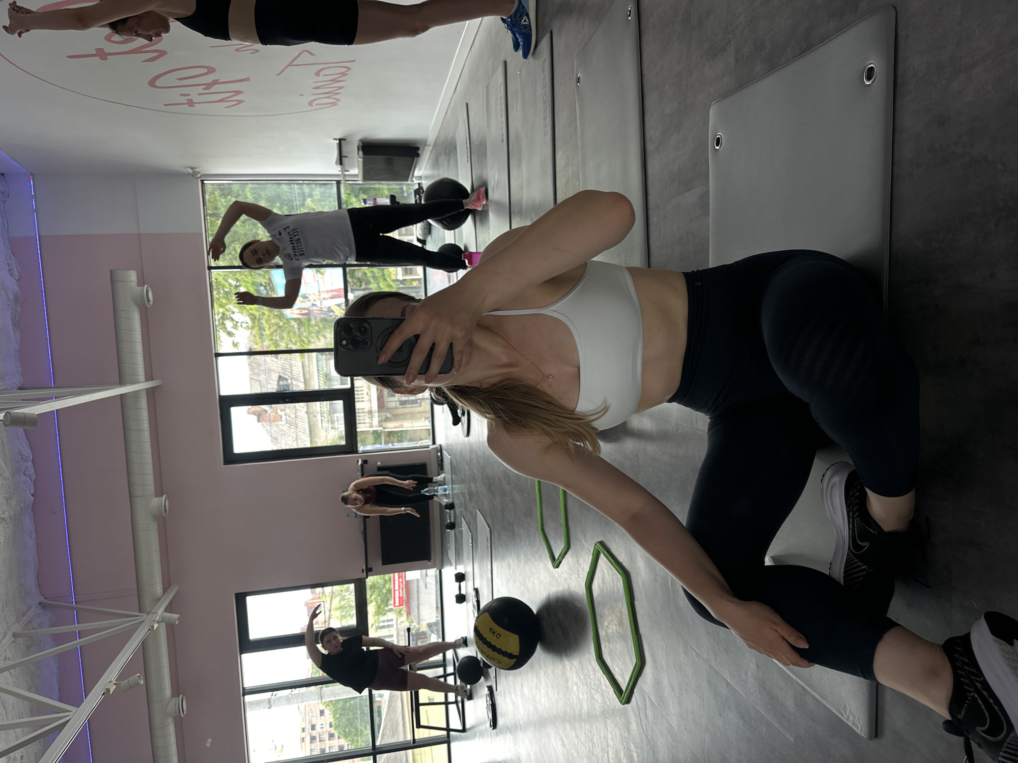This image captures an indoor fitness studio or gym in Ternopil, Ukraine, featuring five individuals engaged in exercise or pre-workout routines. The room has a dark grey floor, pink lower walls, white upper walls, and a white industrial-style ceiling with exposed trusses, ventilation ducts, and track lighting, accented by a subtle purple LED strip. Large, floor-to-ceiling windows span the background, revealing an urban landscape of buildings and greenery, consistent with a city environment in Ternopil.
In the immediate foreground, a woman with light brown hair, wearing a white sports bra, dark leggings, and black Nike sneakers, sits on a grey exercise mat. She holds a dark-colored smartphone (likely an iPhone Pro model) in her right hand, positioned to take a selfie, with her face obscured by the device. Her left arm is bent, resting on her leg.
Further back, from left to right:
1. A man in a black t-shirt and dark shorts performs a side bend or stretch near a window. A black medicine ball (marked "8KG") and two green hexagonal agility rings are on the floor in front of him.
2. A woman with dark hair, wearing a white short-sleeved top and dark leggings, stands with her arms raised, engaged in a warm-up exercise.
3. Behind and to the right of the previous woman, another woman with dark hair, dressed in a dark red sports top and dark leggings, bends forward, possibly stretching or reaching for equipment. A pair of silver dumbbells and a pink water bottle are on the floor nearby.
4. On the far right, a woman with dark hair, clad in a black sports bra and black shorts, stands with her arms raised overhead, facing away from the windows. She wears blue Reebok sneakers.
Several grey exercise mats are laid out across the floor. In the background, to the far left, a black step platform is visible, and on the right wall, a large circular white graphic with pink script lettering (partially visible as "Ternopil") serves as decor. The scene depicts a dynamic fitness environment with various exercise equipment in use.