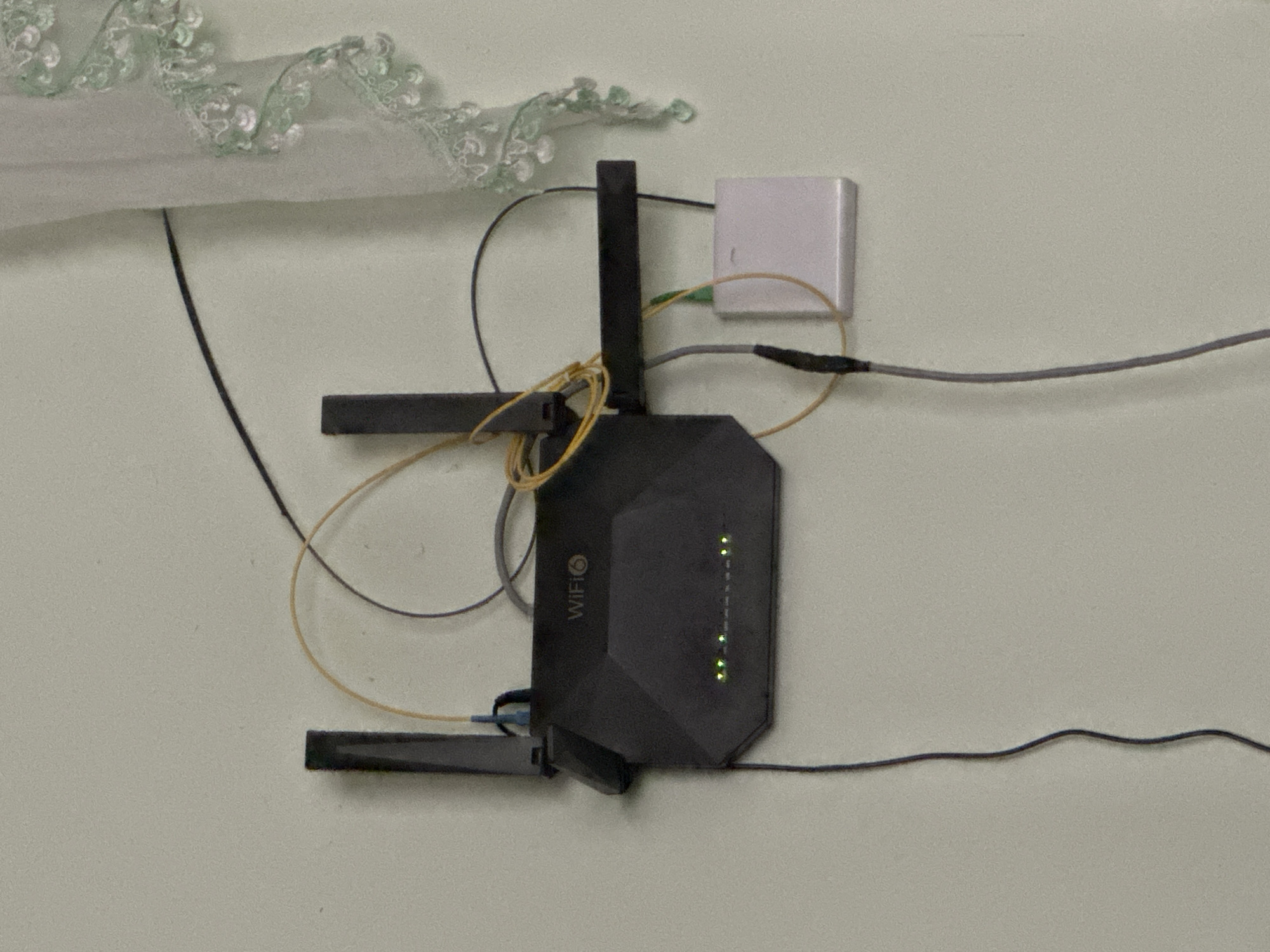 This image depicts a black WiFi router mounted on a pale, textured wall, likely indoors. The router has two antennas and several green indicator lights. A small white box, possibly a modem, is attached to the wall to the right of the router. Various cables, including a yellow one and a grey one, connect the devices and run down the wall. To the right of the router, a sheer white curtain with decorative green trim is partially visible. The overall environment appears to be a domestic setting. There are no people or obvious signs of activity in the image. The location of Alimkent, Uzbekistan is not evident from the visual information in the photograph.