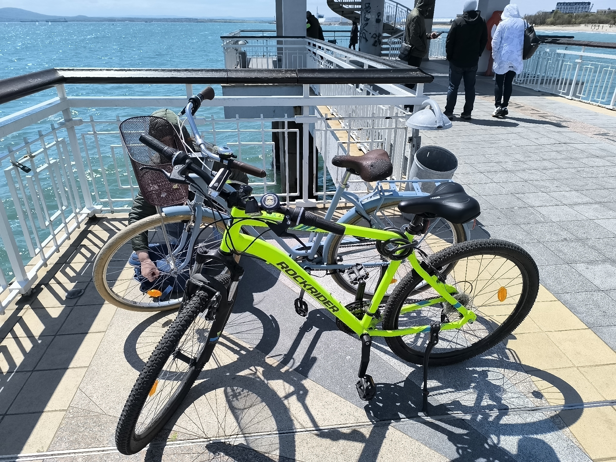 The image depicts two bicycles parked on a paved platform near a body of water. One bicycle is a bright green mountain bike with the brand "ROCKRIDER" on its frame. The other bicycle is a lighter color, possibly light blue or silver, and has a basket on the front and a vintage-style seat. The bicycles are parked near a metal railing, behind which is a blue body of water. In the background, several people are walking on the platform. The sky is clear and bright. Based on the background and architectural details, the location is likely near the sea in Burgas, Bulgaria.