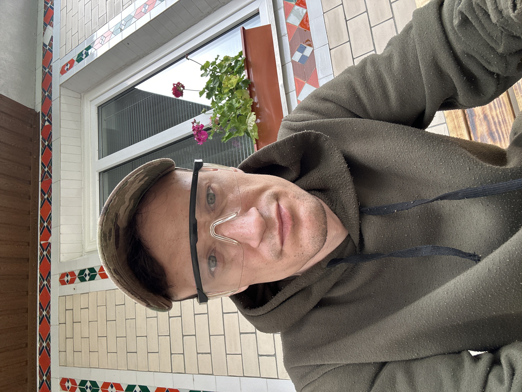 A man wearing a camouflage baseball cap and clear protective eyewear, both of which appear to be in good condition, is pictured from the chest up. He is wearing a dark olive green hoodie with the hood up, which is pilled and appears worn. He is seated outside, indicated by a wooden surface beneath him. Behind him, a brick wall with decorative tile accents runs along the side of a building. A window with blinds and a planter box containing two pink flowers is visible above the tilework. The building's facade is made of light beige bricks, and a brown wooden soffit is visible at the top of the frame. The scene is consistent with an outdoor setting in a residential area, potentially Makiv, Ukraine, given the architectural style.