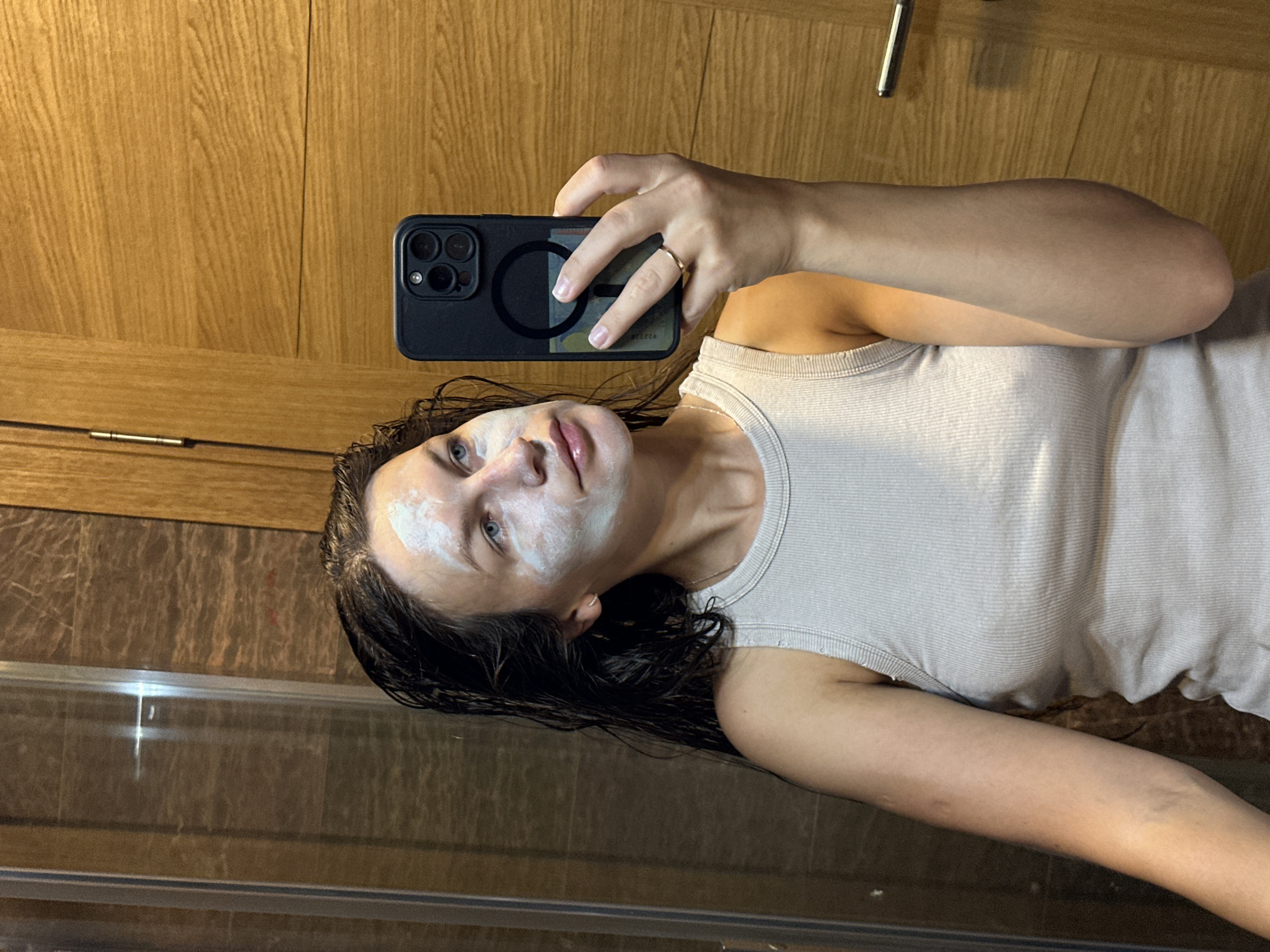 A woman with wet, dark hair and a white face mask applied to her face is taking a mirror selfie in a bathroom. She is wearing a light gray ribbed tank top and a thin gold ring on her right ring finger. She is holding a black smartphone in her right hand. The bathroom features dark tiled walls and light wood paneling. A glass shower door is visible to the left, and a wooden door with a silver handle is to the right. The overall lighting suggests it is indoors, possibly evening or in a room with artificial light. The location is identified as Donji Pobori, Montenegro.
