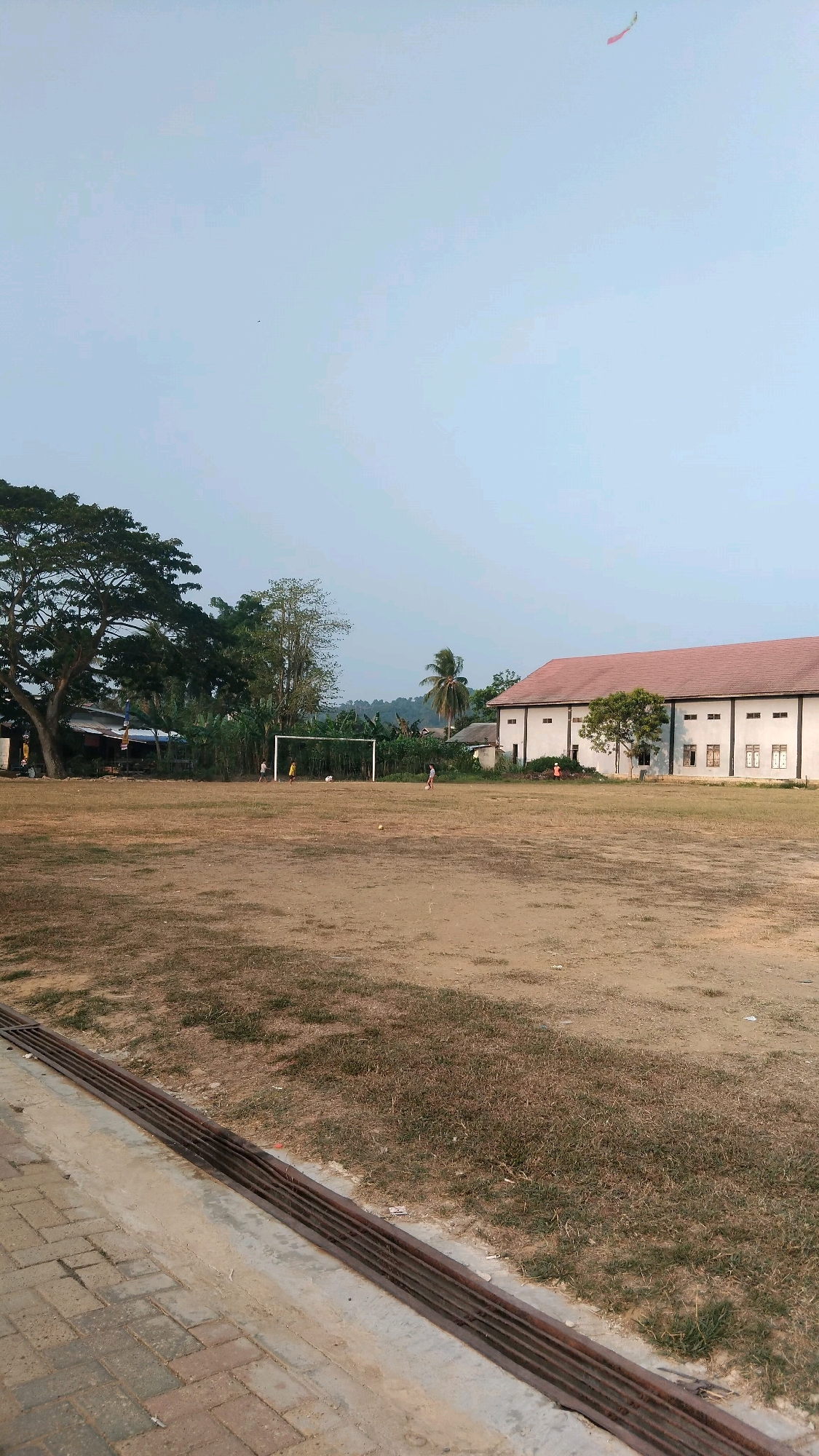 The image depicts an outdoor scene at what appears to be a community soccer field in Manunggal Jaya, Indonesia, during daylight hours. The field itself is predominantly dry, with patches of sparse grass and exposed soil. In the background, a large, multi-story building with a red-tiled roof and white exterior walls stands, featuring numerous windows. To the left of the building, several trees are visible, including a large, leafy one and a palm tree. A soccer goal is positioned on the field, and a few individuals, appearing to be children or young adults, are scattered across the playing area, engaged in activities that suggest a casual game of soccer. Two soccer balls are also discernible on the field. In the sky, a small kite is visible flying in the upper right portion of the frame. The foreground shows a paved walkway made of brick pavers adjacent to a drainage grate. The overall environment suggests a warm, possibly tropical climate.