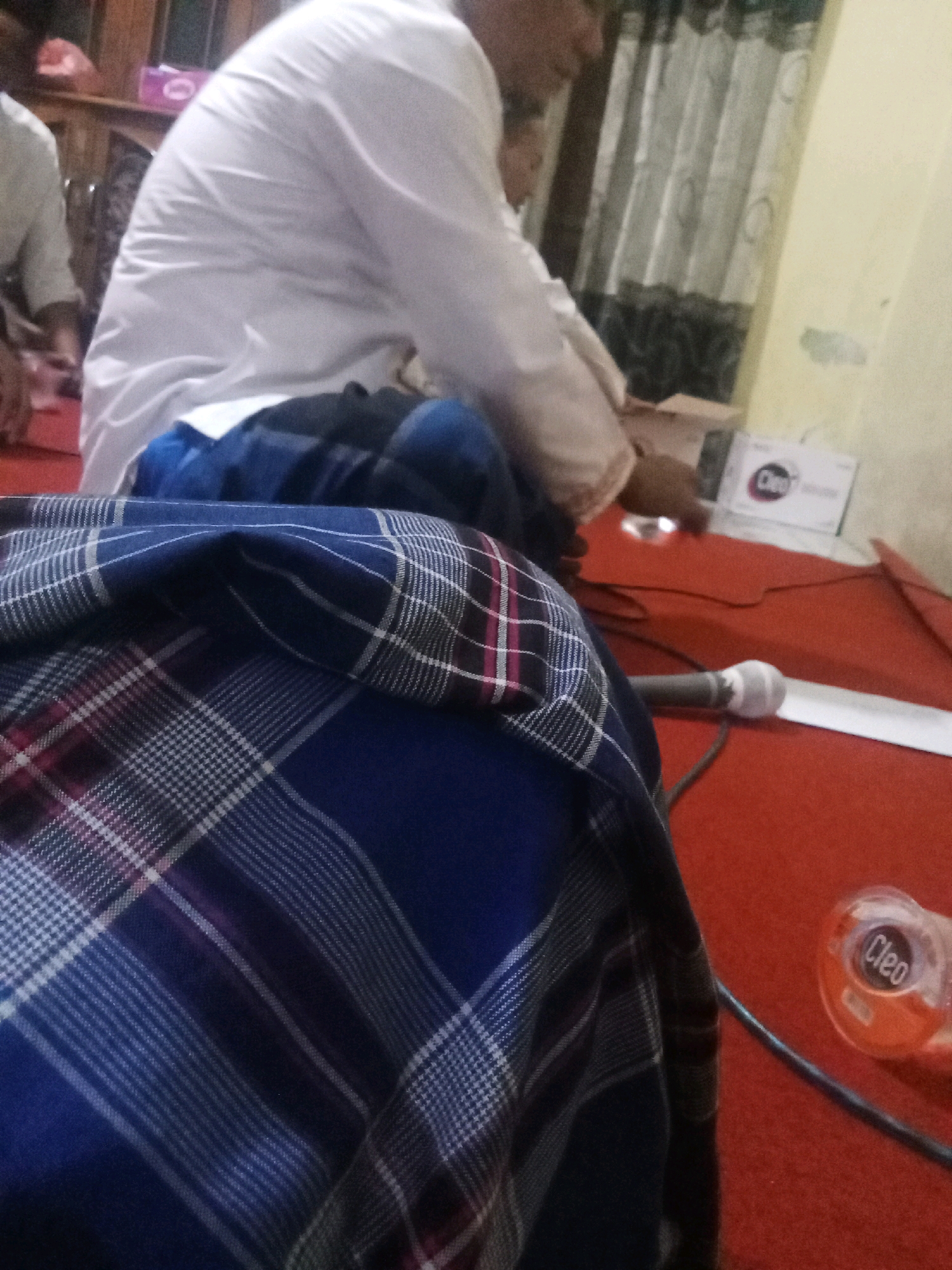 In Keplaksari, Indonesia, the image depicts an indoor scene with a red carpeted floor. In the foreground, a blue and white checkered fabric is draped, obscuring much of the lower frame. Behind this fabric, a person wearing a white garment, possibly a long shirt or robe, is seated. Their face is partially visible and appears to be looking down. To the right of the person, a microphone on a stand is visible, and further to the right, a small orange bottle with a label that reads "Cleo" is present. In the background, curtains hang on a wall. The overall setting appears to be a casual gathering or event.