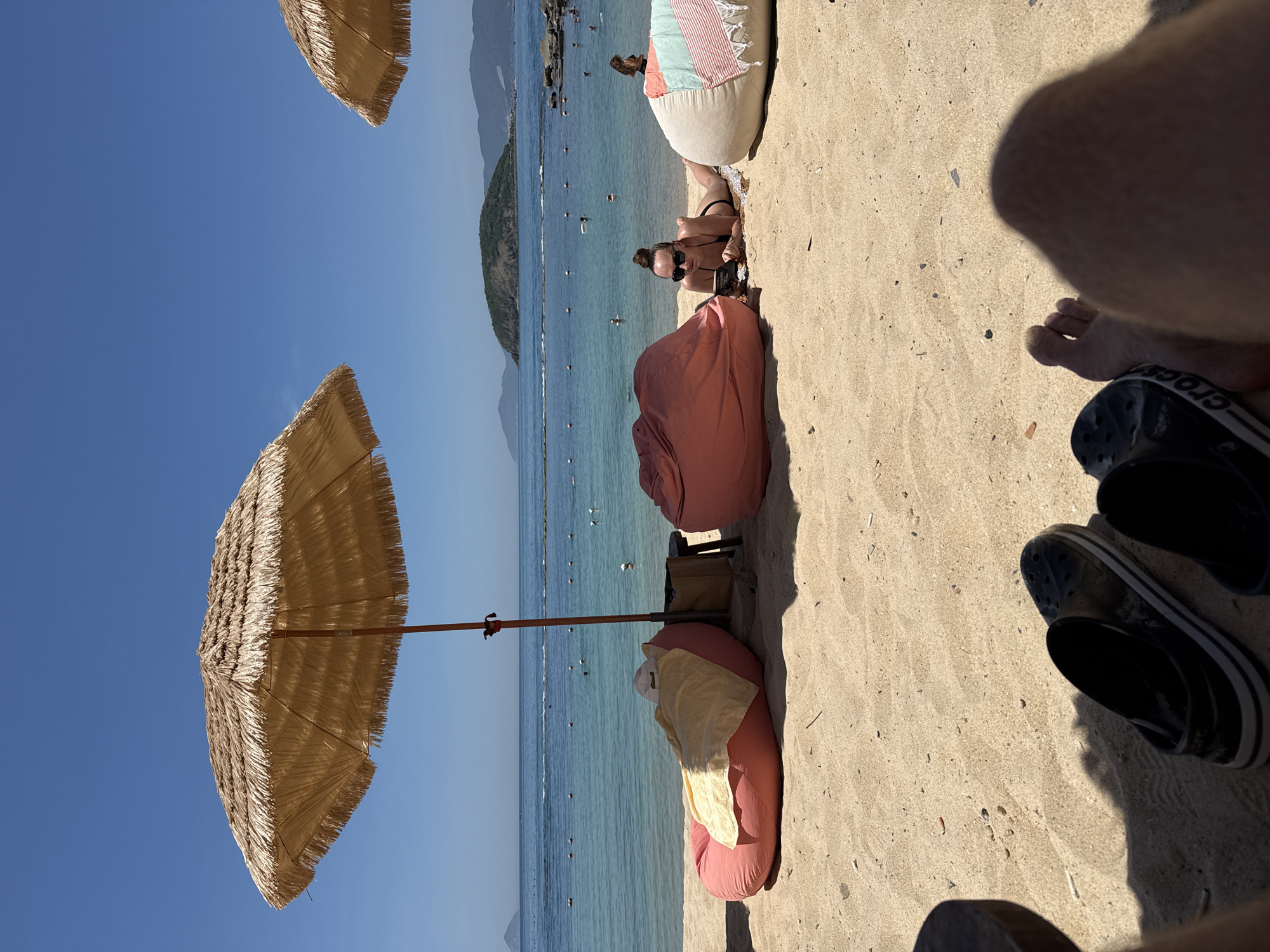 The image depicts a sunny day at a beach in Nha Trang, Vietnam. The scene is oriented vertically, showing a sandy beach with the ocean in the background. Several straw umbrellas are visible, one in the foreground providing shade for two pink beanbag chairs, one of which has a yellow towel draped over it. In the mid-ground, a woman in a black bikini and sunglasses is lying on a beanbag chair, holding a phone. More beanbag chairs and beach umbrellas are scattered along the sand. The turquoise water is calm, with numerous small buoys and a few people swimming or wading in the distance. Mountains form a backdrop to the coastline. The lighting suggests it is daytime, likely mid-morning or afternoon, with clear skies and warm weather. The overall impression is one of relaxation and leisure at a tropical beach resort. There is no visible text in the image.