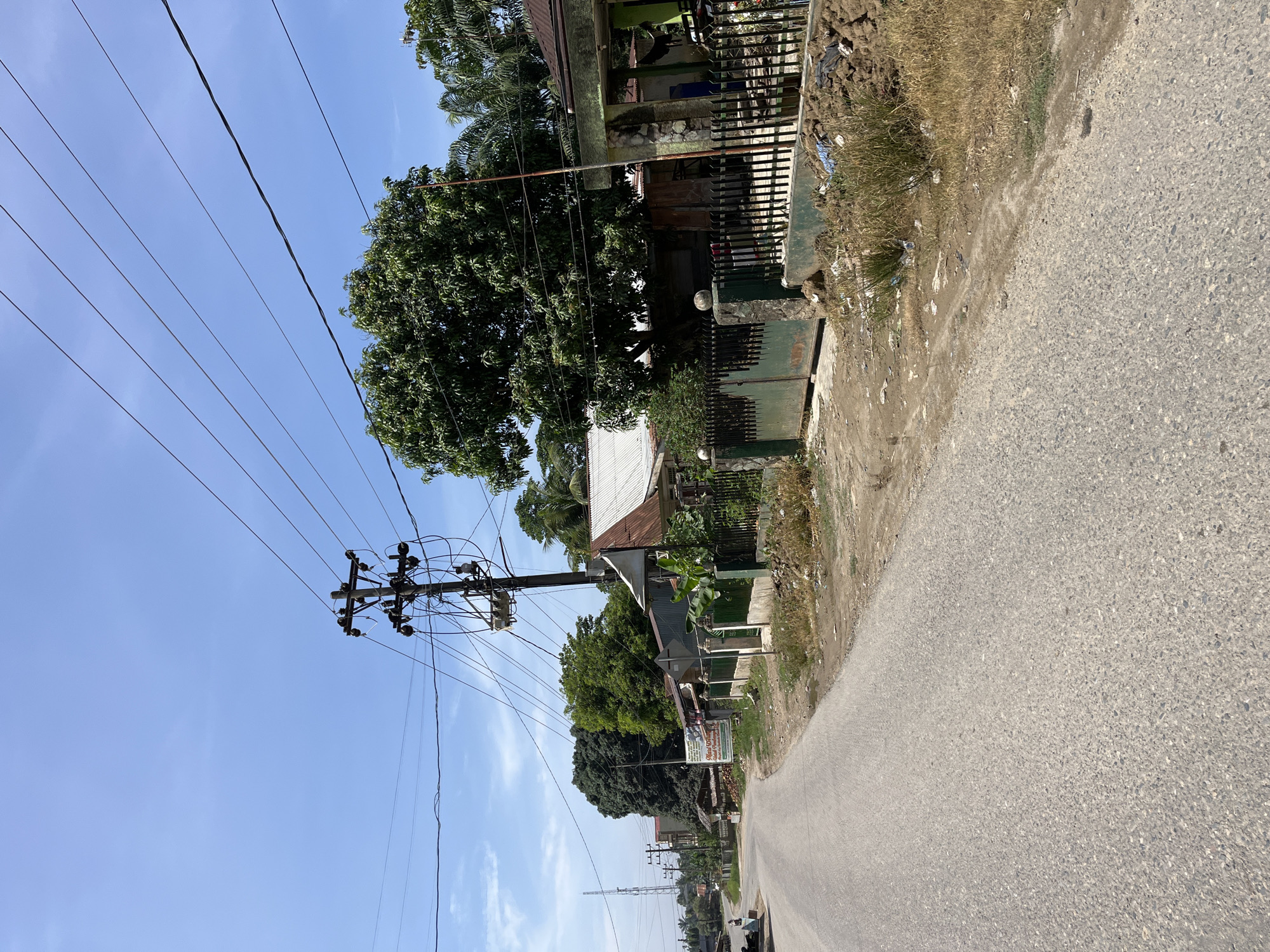 The image depicts a street scene in Landuh, Indonesia. The foreground shows a paved road curving to the left. On the right side of the road, there is a dirt shoulder with dry grass and debris. Beyond the shoulder, a green metal gate with stone pillars leads to a property with houses, fences, and lush green trees. Overhead, utility poles with numerous electrical wires traverse the sky, which is clear blue with some scattered white clouds. In the distance, a tall tower is visible. Several people are seen on the left side of the road, near some buildings. The environment appears to be a rural or semi-urban area.