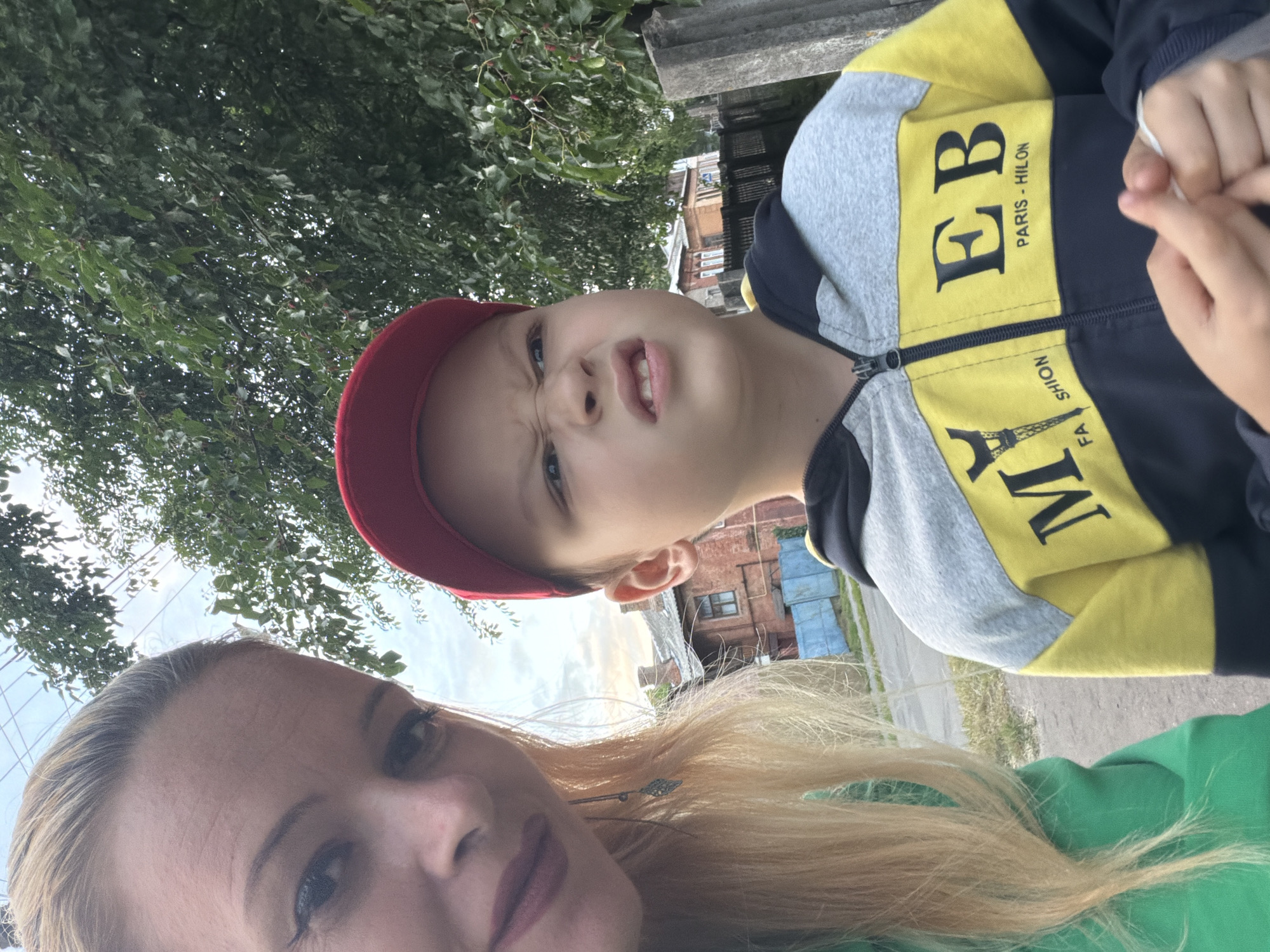In this close-up, outdoor selfie taken in Chernihiv, Ukraine, a woman with blonde hair and makeup is on the left, and a young boy with a red cap is on the right. The woman is wearing a green hoodie, and the boy is wearing a grey, yellow, and navy blue zip-up hoodie. The boy is making a grimace, and his hands are visible in the foreground, holding a white object. Behind them, a tree-lined street is visible with houses and a wooden fence. The sky is overcast.