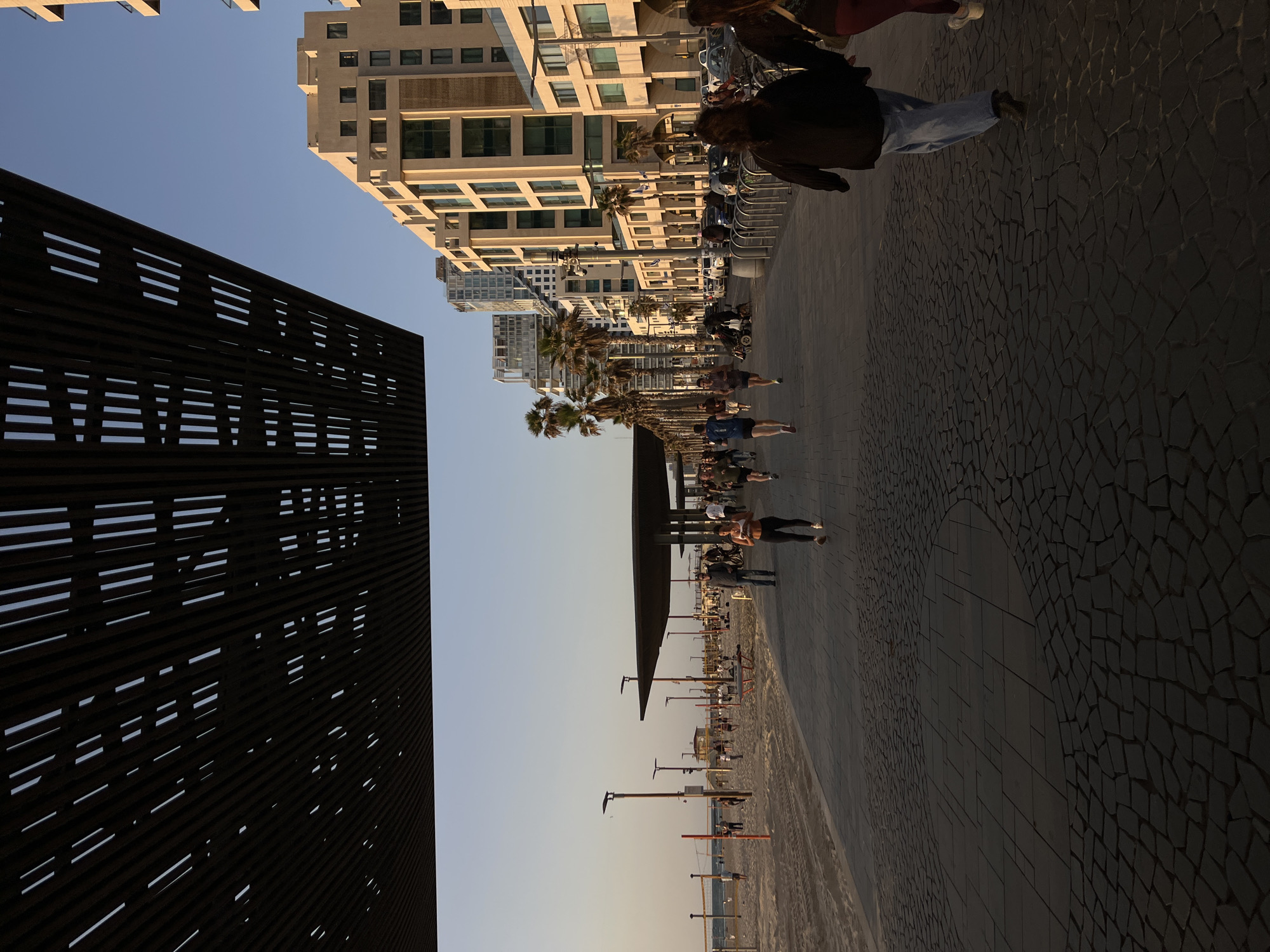 The image depicts a Tel Aviv beachfront scene. A modern, perforated, dark architectural structure is on the left. To the right is a paved walkway lined with palm trees, leading to a sandy beach. People are walking and congregating along the walkway and on the beach. Buildings, including modern apartment blocks, rise in the background. The time of day appears to be late afternoon or early evening, indicated by the angle and color of the light.