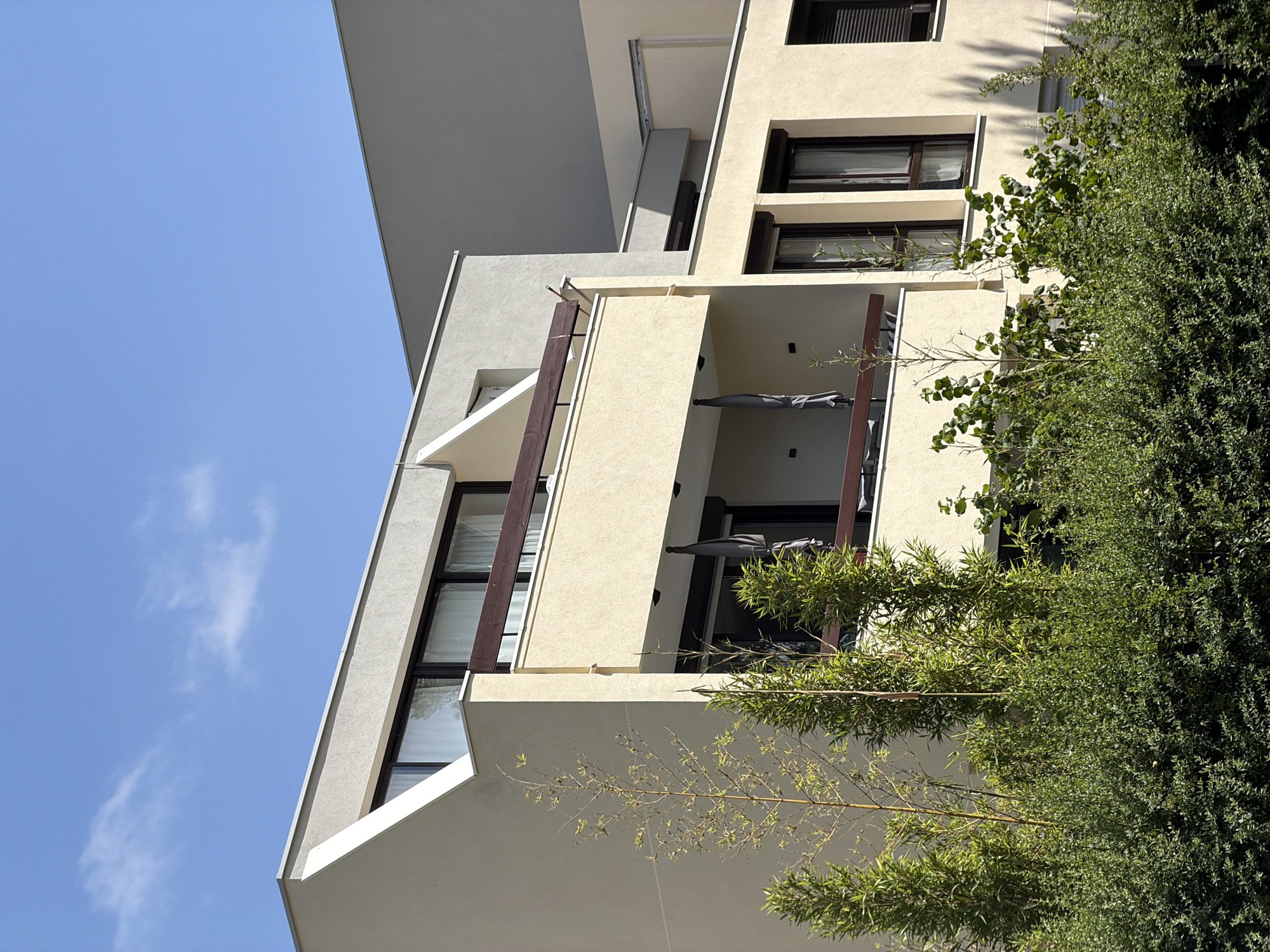 This is a low-angle, vertical shot of a modern building with balconies and large windows, partially obscured by lush green foliage. The building's facade is a mix of light cream and grey concrete. Several balconies are visible, some with folded grey umbrellas. The windows are dark-framed and reflect the bright blue sky and some white clouds. Thin, wooden beams run vertically along some of the balcony edges. Thin branches with small green leaves, possibly bamboo, extend from the right side of the frame and trail across the lower part of the building. The overall impression is of a contemporary residential building integrated with nature. The scene is illuminated by bright sunlight, suggesting it is daytime with clear weather. No people or text are visible. The context provided places the location in Évry-Courcouronnes, France.