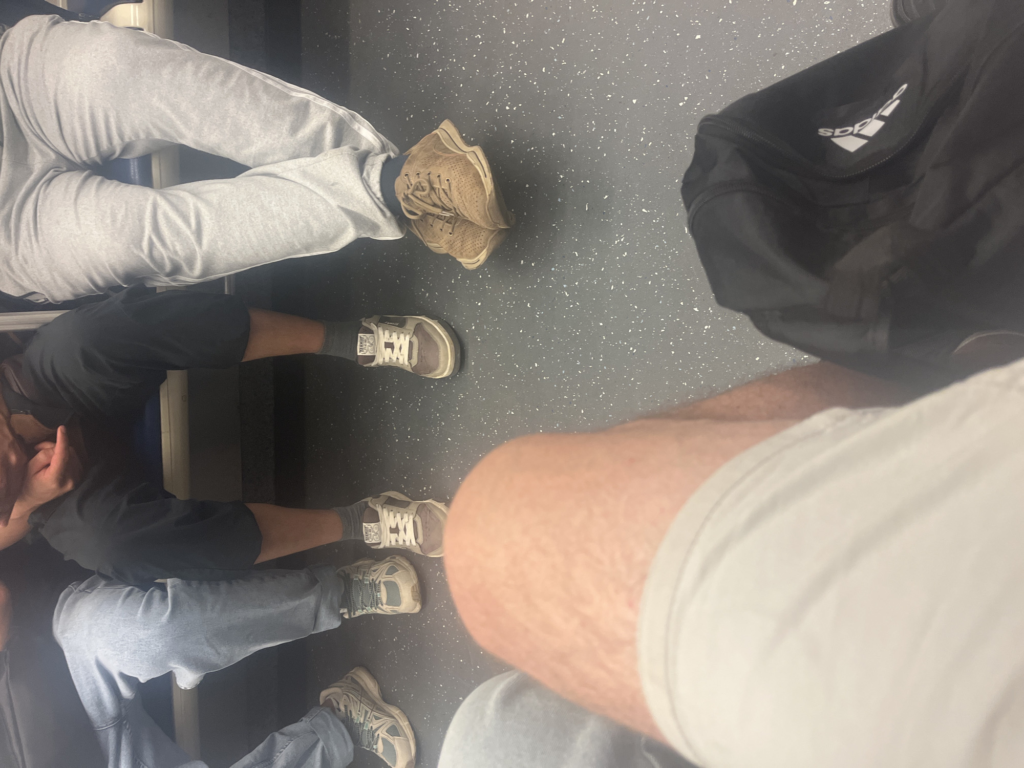 The image depicts a low-angle view of several individuals' legs and feet, seated closely together, likely on public transportation. The flooring is a speckled grey surface. On the left, one person wears light blue jeans and white sneakers with grey accents. Next to them, another person is in dark pants and grey socks with brown and white sneakers. Further to the right, a person in light grey sweatpants with white stripes down the side wears dark socks and brown, textured sneakers. Another individual's legs are visible in the foreground, wearing white shorts. A black Adidas bag is partially visible in the bottom right corner. The setting is indoors, with a cool, artificial light. There is no visible indication of Moscow, Russia in the image.