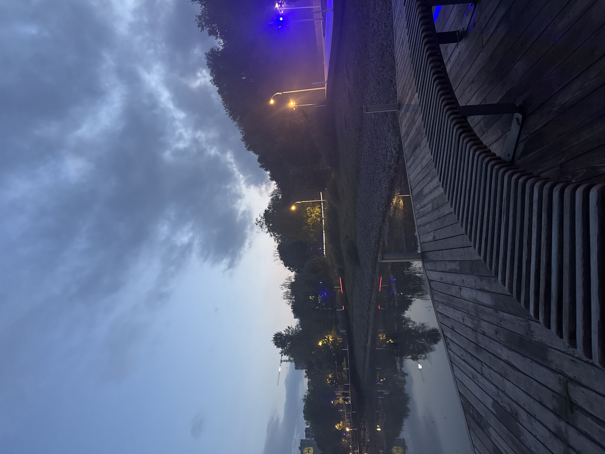 The image shows a tranquil park setting at dusk, likely located in Ufa, Russia. A wooden pier or boardwalk extends into a calm body of water, with a modern, slatted bench positioned in the foreground. The water reflects the dim lights of the surrounding area and the overcast sky. Beyond the water, trees line the bank, illuminated by streetlights and some vibrant blue lighting further in the background, possibly from a performance or event. In the distance, city buildings and a construction crane are visible against the twilight sky, indicating an urban or semi-urban environment. The overall atmosphere is serene and slightly mysterious due to the low light conditions.