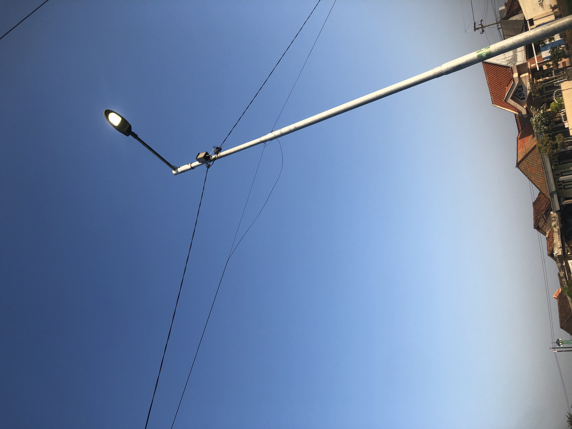 This image depicts a street scene in Kalitengah, Indonesia. A tall, light-colored streetlamp with its light illuminated stands prominently against a clear, bright blue sky. Several dark power lines crisscross the upper portion of the frame, some appearing to be connected to the streetlamp pole. In the lower part of the image, a row of residential buildings with tiled roofs is visible, suggesting a suburban or semi-urban environment. The architecture of the houses appears typical of the region, with some featuring peaked roofs and decorative elements. The overall lighting suggests it is daytime, possibly late afternoon or early morning, given the clarity of the sky and the illumination of the streetlamp. There are no people or animals visible in the frame.