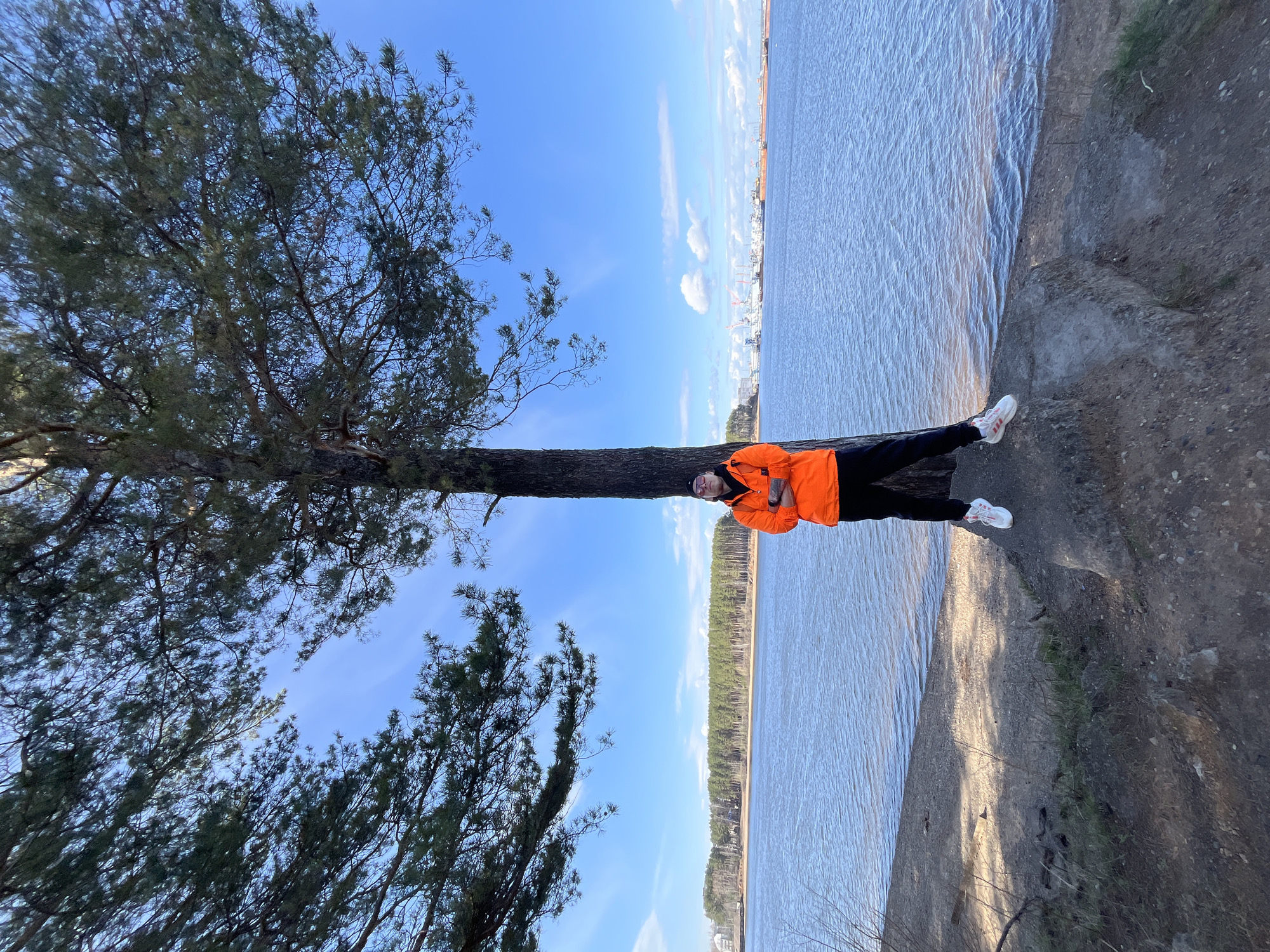The image, taken in Chaikovsky, Russia, depicts a single person standing outdoors on a sunny day. The individual is positioned against the trunk of a large, tall pine tree, which dominates the left side of the frame with its dark green foliage.
The person is wearing a bright orange jacket, black pants, and white sneakers. They have a dark beanie on their head and appear to be wearing eyeglasses or sunglasses. Their arms are crossed as they look towards the camera.
To the right of the person and the tree, a wide body of water, likely a river or large lake, stretches across the mid-ground to the horizon. The water's surface shows subtle ripples and reflects the clear blue sky. The person is standing on a textured bank of dirt and sparse vegetation that borders the water.
In the far background, beyond the water, a dense treeline marks the opposite shore. Above this distant treeline, faint outlines of what appear to be industrial structures or cranes are visible, suggesting a distant urban or port area. The sky above is a brilliant, clear blue with a few scattered wispy clouds, indicating bright and pleasant weather. There is no visible text in the image.