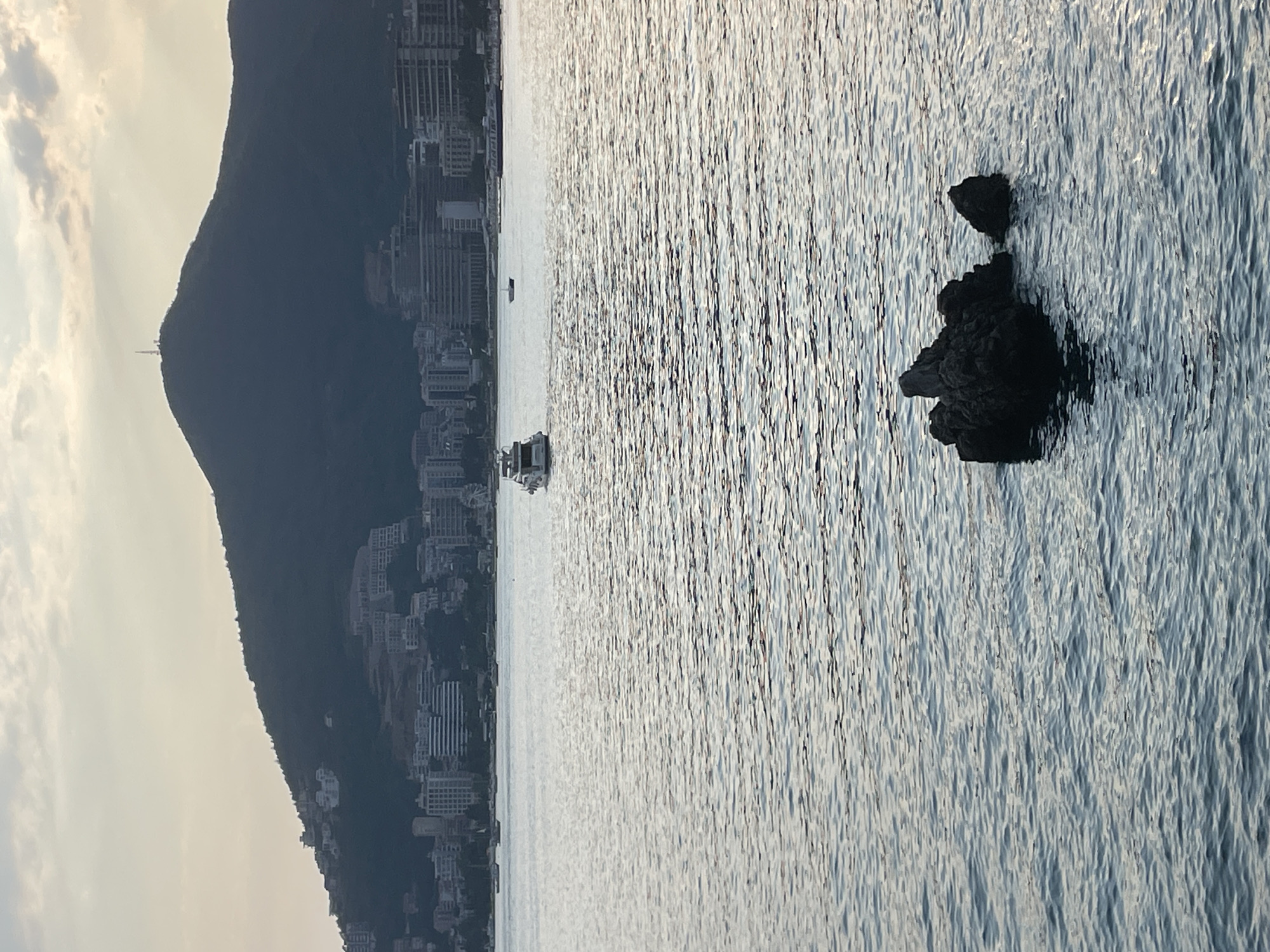 The image depicts a coastal scene in Budva, Montenegro, looking towards the mountains and the sea. In the foreground, a cluster of dark rocks emerges from the rippling blue water. Further out, a white motor yacht is visible, leaving a wake as it moves across the water. In the background, the city of Budva is spread out along the coastline, with numerous buildings clustered at the base of a large, dark mountain. The mountain is capped with a communication tower and appears to be covered in vegetation. The sky above is a pale, hazy yellow, with soft clouds, suggesting either sunrise or sunset. The overall impression is one of a serene, late-afternoon or early-morning view of the Adriatic coast.