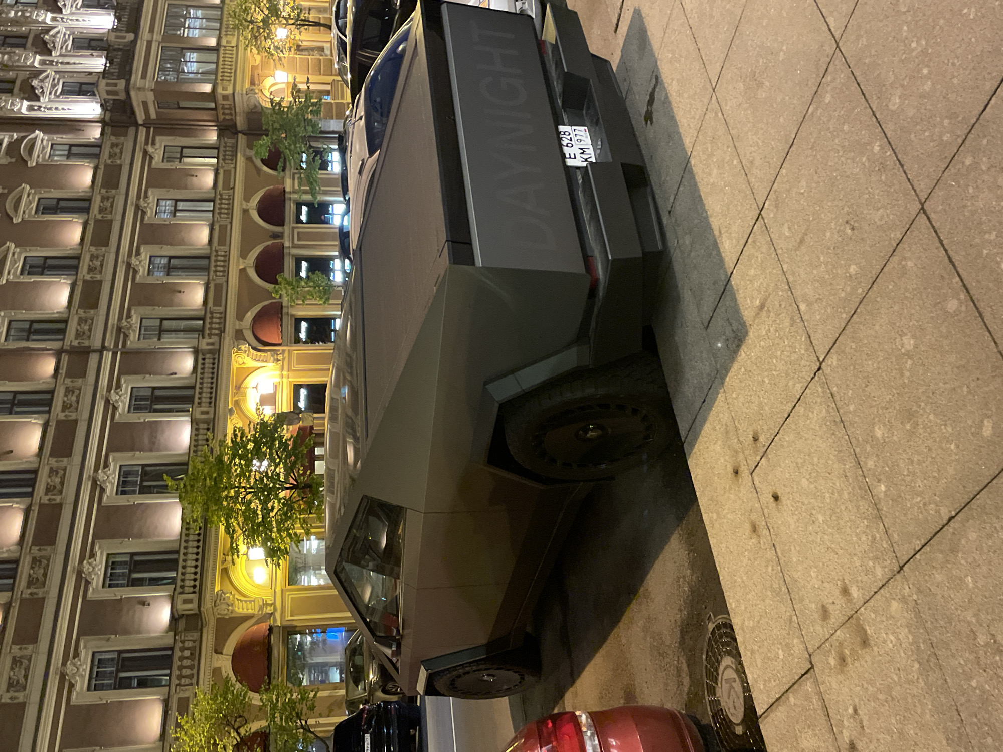 The image shows a modified Tesla Cybertruck parked on a street in Saint Petersburg, Russia. The vehicle has the word "DAYNIGHT" printed vertically on the rear. The license plate is E 628 KM 977. The background includes a building with ornate architecture. The street surface is paved with stone tiles. Other vehicles are partially visible nearby. The time of day appears to be evening or night, indicated by the building's illuminated windows and street lighting.