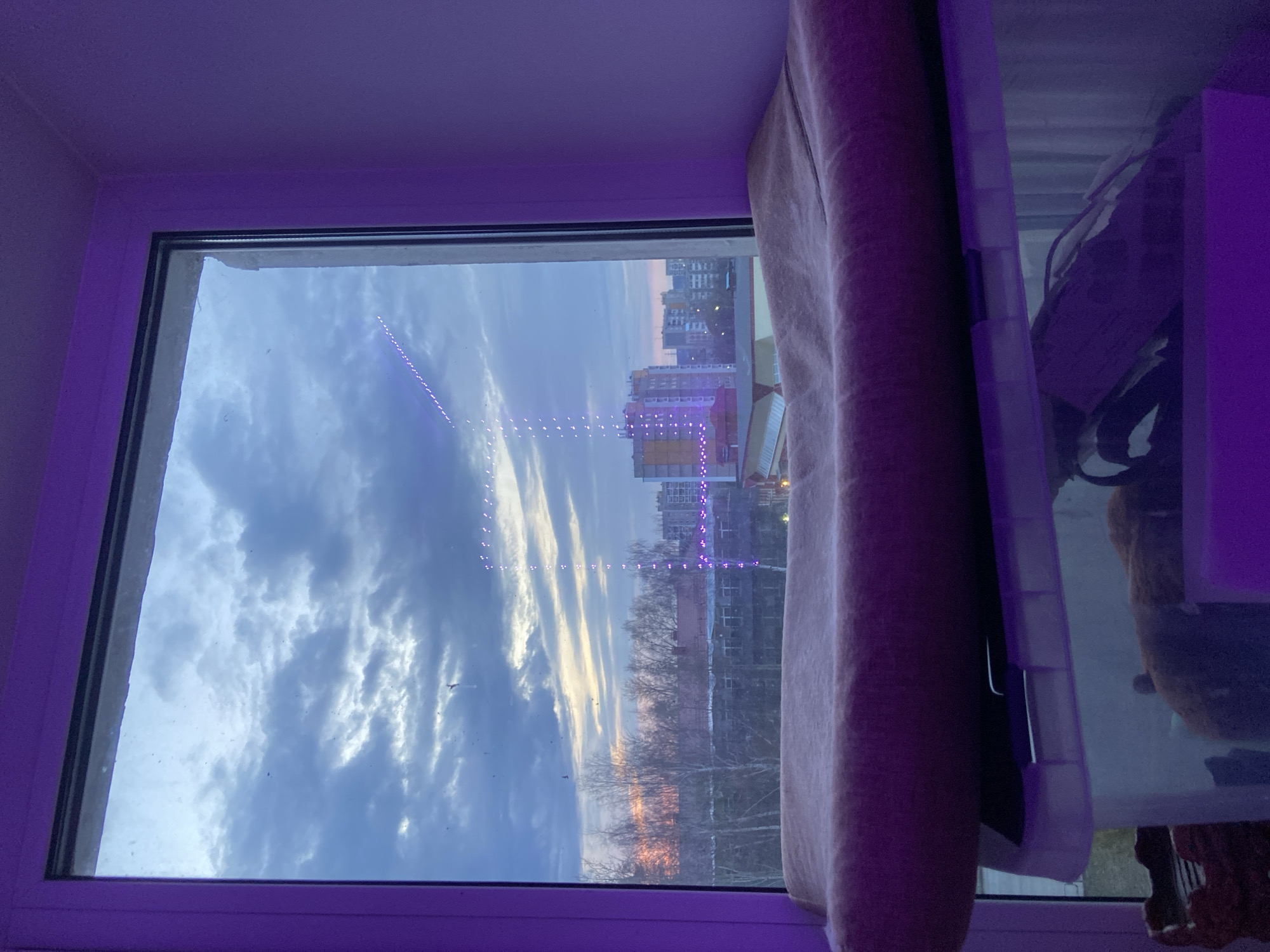 A view from a window shows a cityscape under a partially cloudy sky. In the foreground is a white window frame. The frame is illuminated by purple light. Through the window, the sky is visible with a mix of blue and white clouds. The sun is setting on the horizon, casting an orange glow. In the distance, buildings can be seen, including one brightly lit with purple lights. Below the buildings, there is an expanse of trees. A curtain is to the right of the window. A clear plastic container with contents, and additional objects are visible to the right of the curtain.