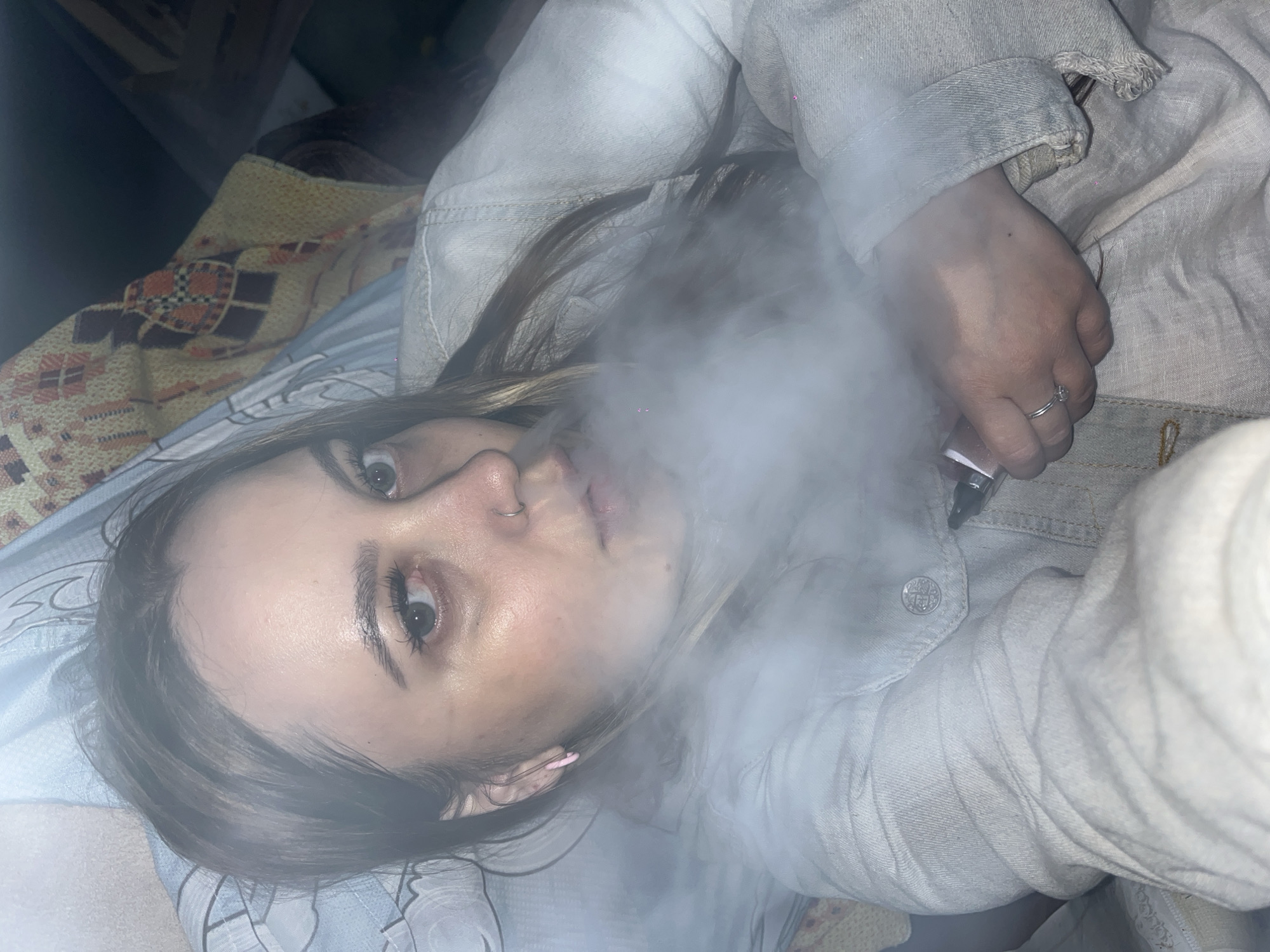 A young woman is lying down, exhaling a large cloud of vapor from a vaping device held in her right hand. She has fair skin, light brown hair, and a nose ring. She is wearing a light-colored denim jacket. The background includes a bed with patterned blankets and sheets. The location of Novopokrovskaya, Russia is not discernible from the image.