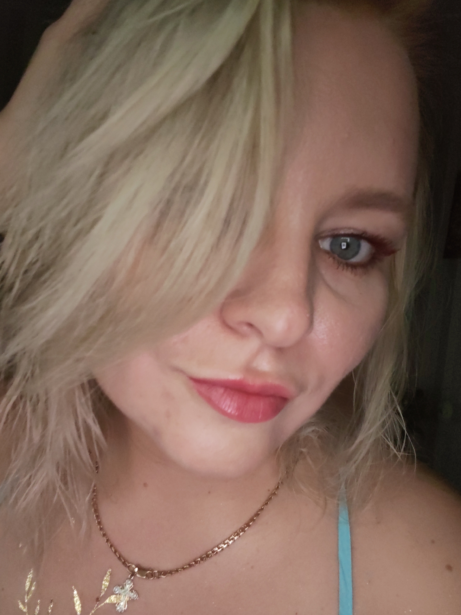 A close-up, shoulder-up, indoor portrait of a young Caucasian woman with blonde hair and blue eyes. She is wearing a rose gold chain necklace with a small, ornate cross pendant. Her lips are painted with a glossy pink lipstick, and she has a hint of red eyeliner on her upper eyelids. Her left hand is partially visible, touching her hair on the top left of the frame. The lighting is dim, with the primary light source appearing to be from the front and slightly from the right, illuminating her face and necklace. There is faint gold body art, resembling leaves, on her chest and shoulder. The background is dark and indistinct. The image is likely a selfie taken in a residential setting in Izhevsk, Russia.